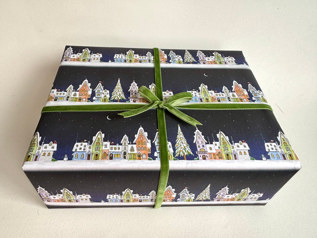 A rectangular sample gift box showing Christmas Village wrapping paper with a colorful houses and tree pattern, tied with a green ribbon and bow.
