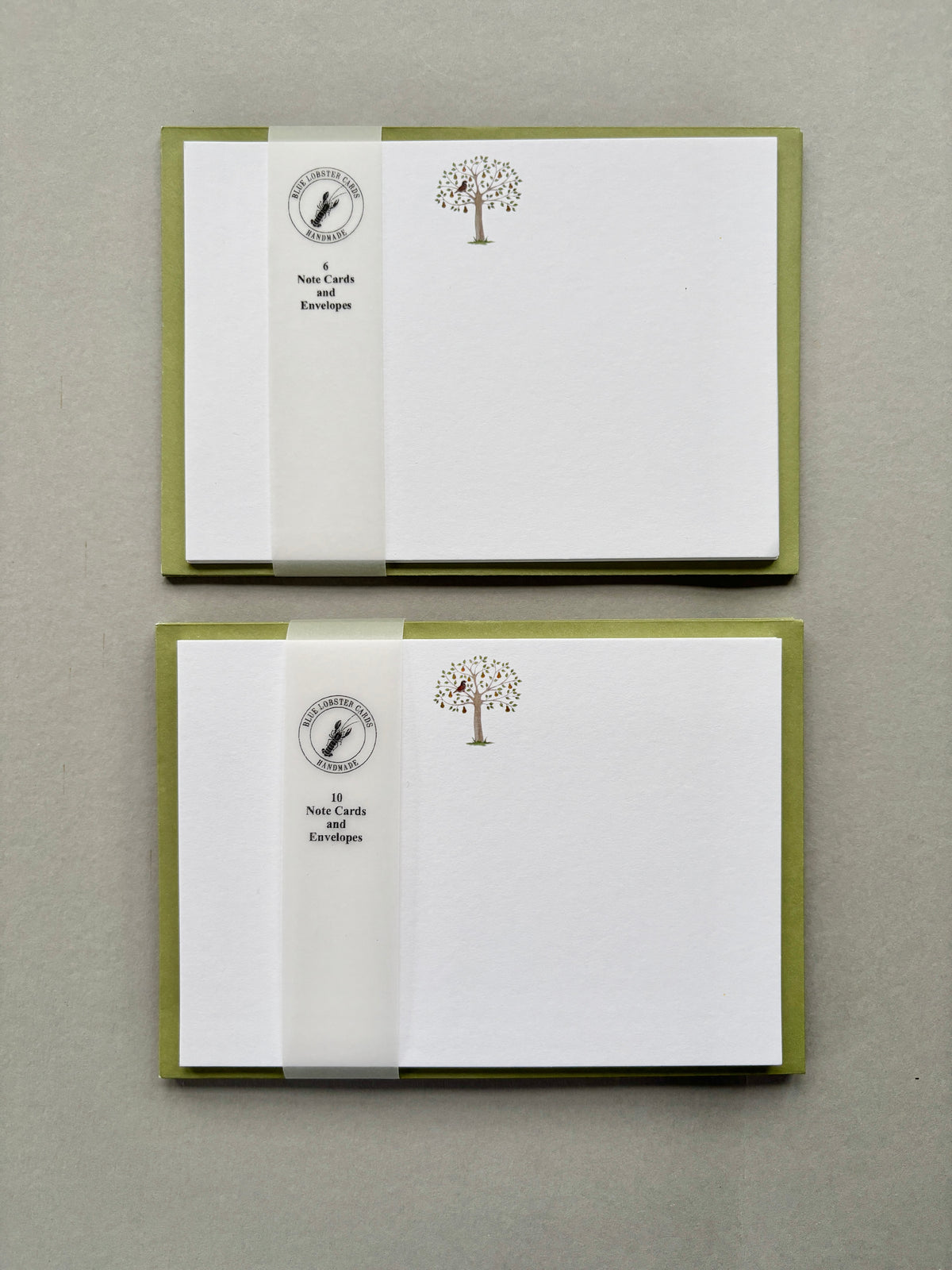 Two stacked packs of 6 and 10 Christmas stationery cards with olive green envelopes, featuring a Partridge in a Pear Tree and Blue Lobster Cards translucent belly band packaging, displayed on a neutral gray background.