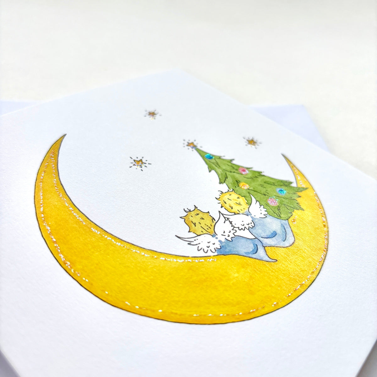 A square white greeting card featuring a hand-drawn illustration of a yellow crescent moon, two small angels, and a decorated Christmas tree, photographed at an angle to show glitter and fine quality matt cardstock
