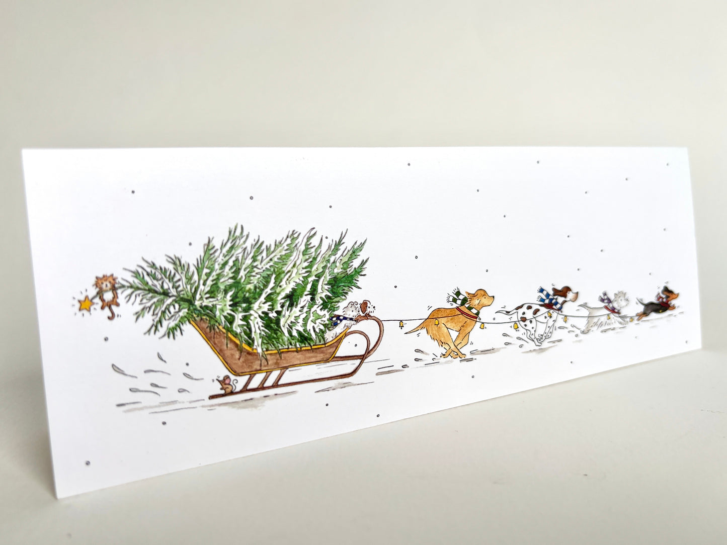 Angle view of fun dog charity Christmas card featuring own original watercolour illustration of dog breeds pulling a snowy Christmas tree through the snow on a sledge