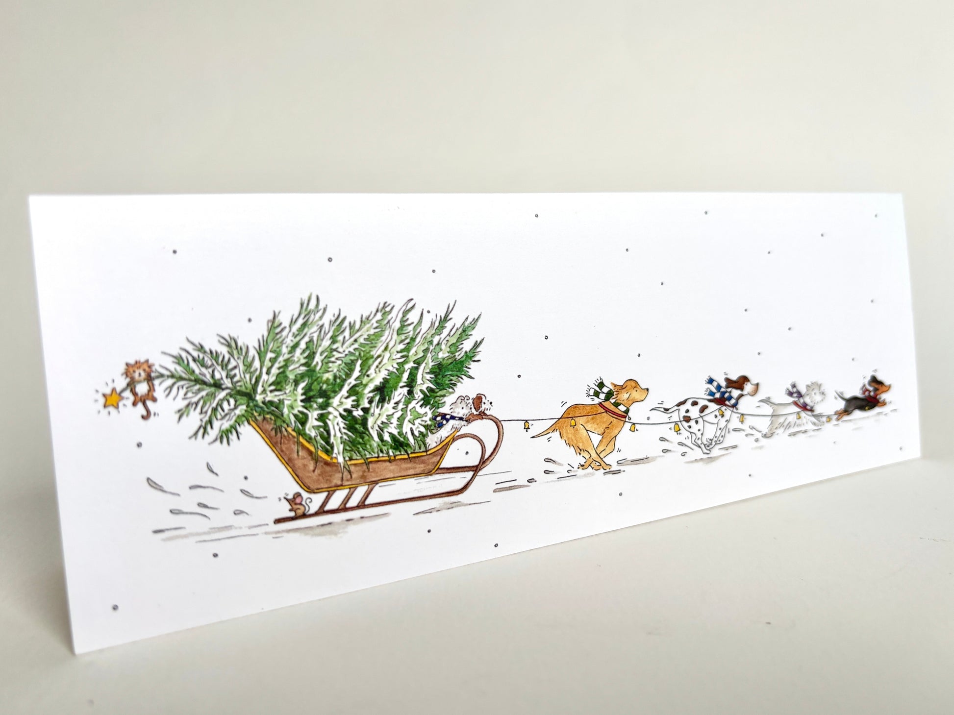 Angle view of fun dog charity Christmas card featuring own original watercolour illustration of dog breeds pulling a snowy Christmas tree through the snow on a sledge