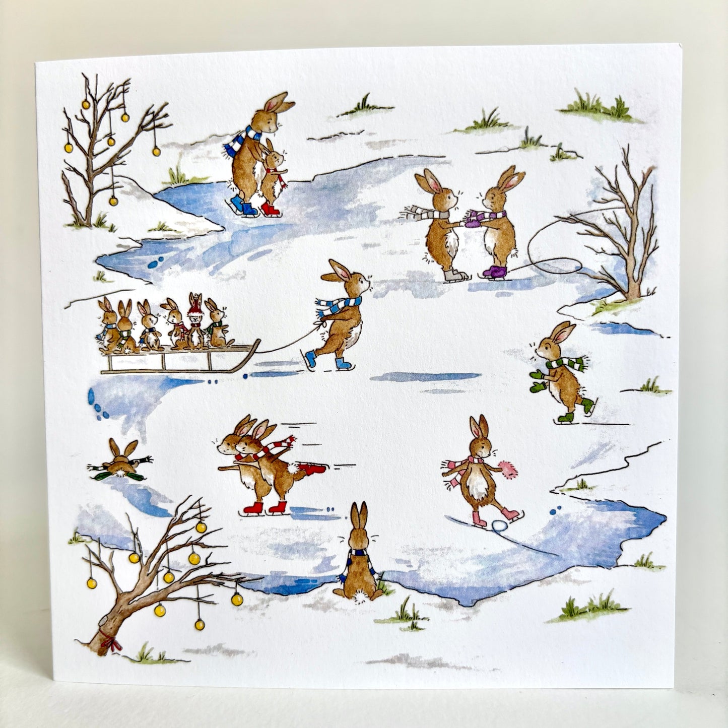 Fine quality square charity Christmas card featuring an original watercolour winter illustration of rabbits ice skating, sledding, and walking through snowy ground, with bare trees and small patches of grass. The card is photographed standing upright against an ivory background.
