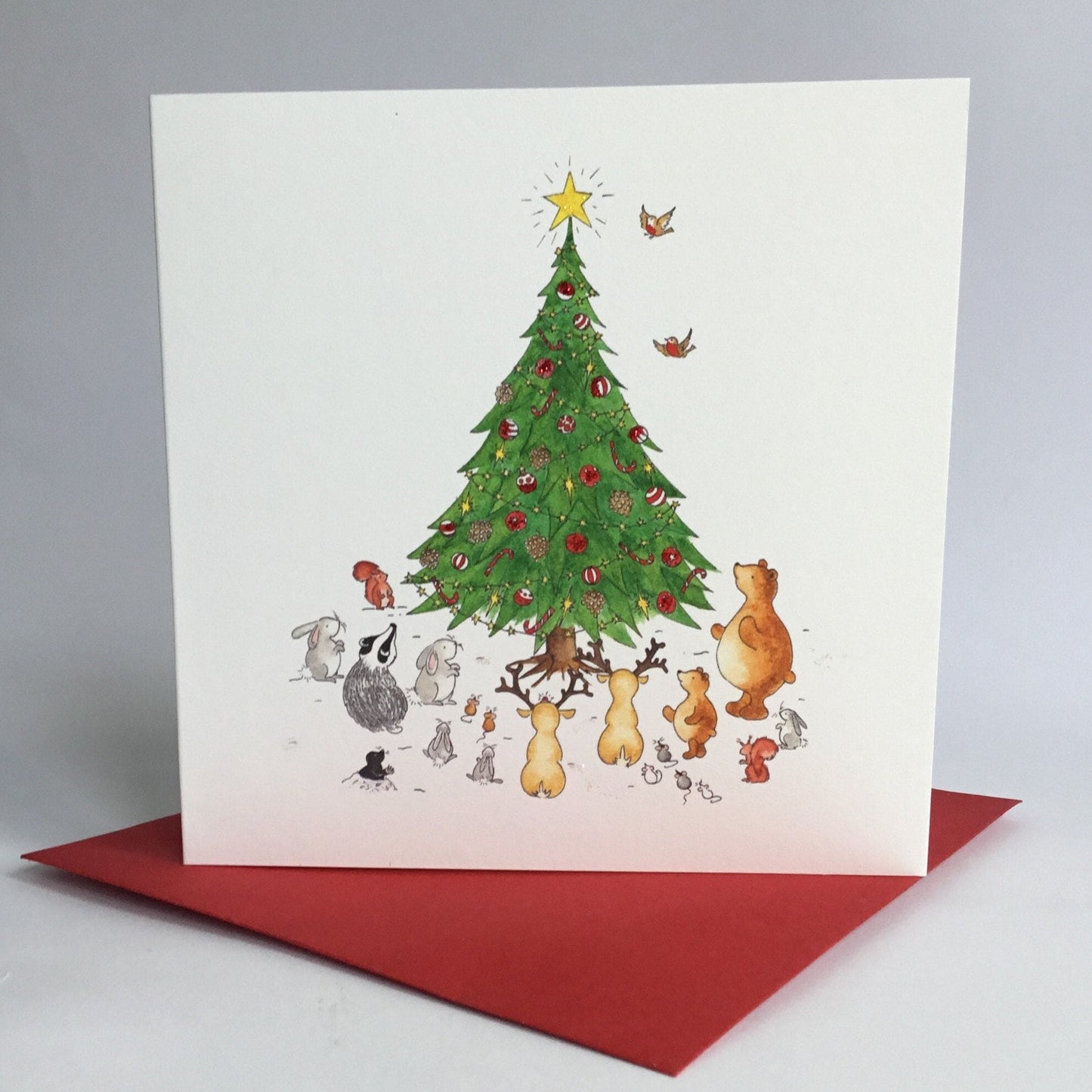 Square christmas card featuring woodland animals surrounding a large tree decoarted with baubles and Christmas star, and standing on a matching red envelope