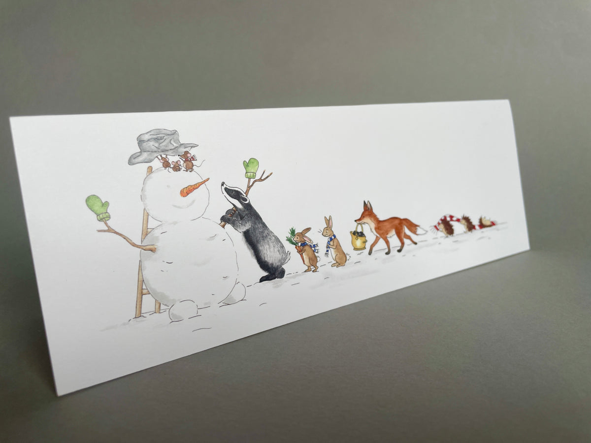Angled front view of a charity Christmas card showing a badger helping woodland animals build a snowman designed by Blue Lobster Cards