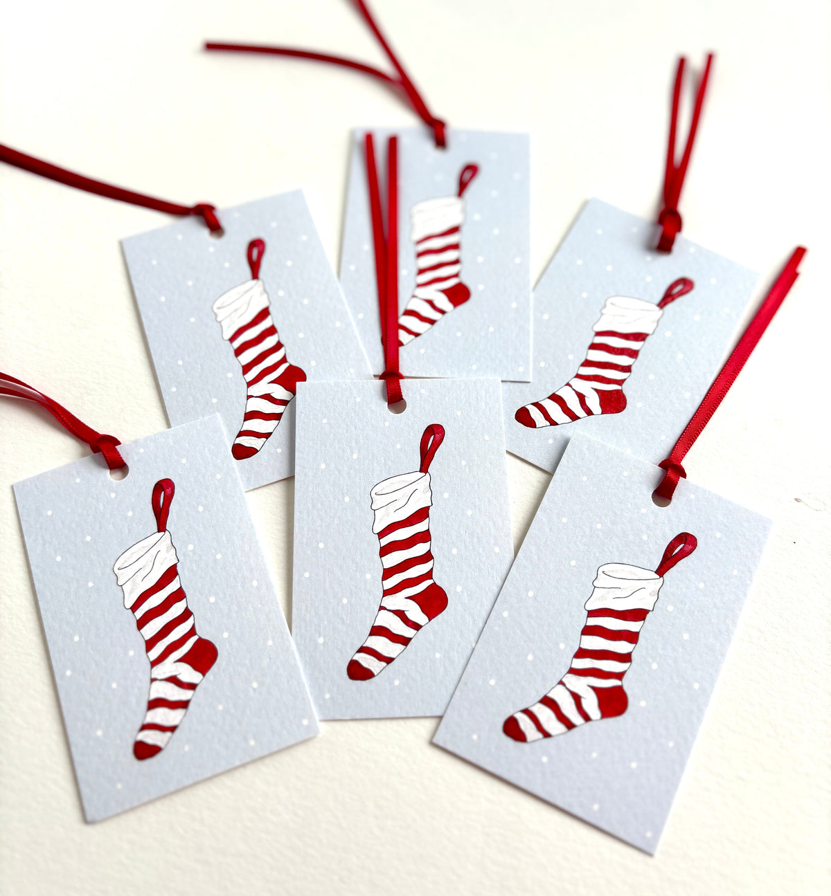 et of 6 rectangular gift tags with hand-tied red satin ribbons, printed with a red-and-white striped Christmas stocking on an ice-blue background with falling snow.