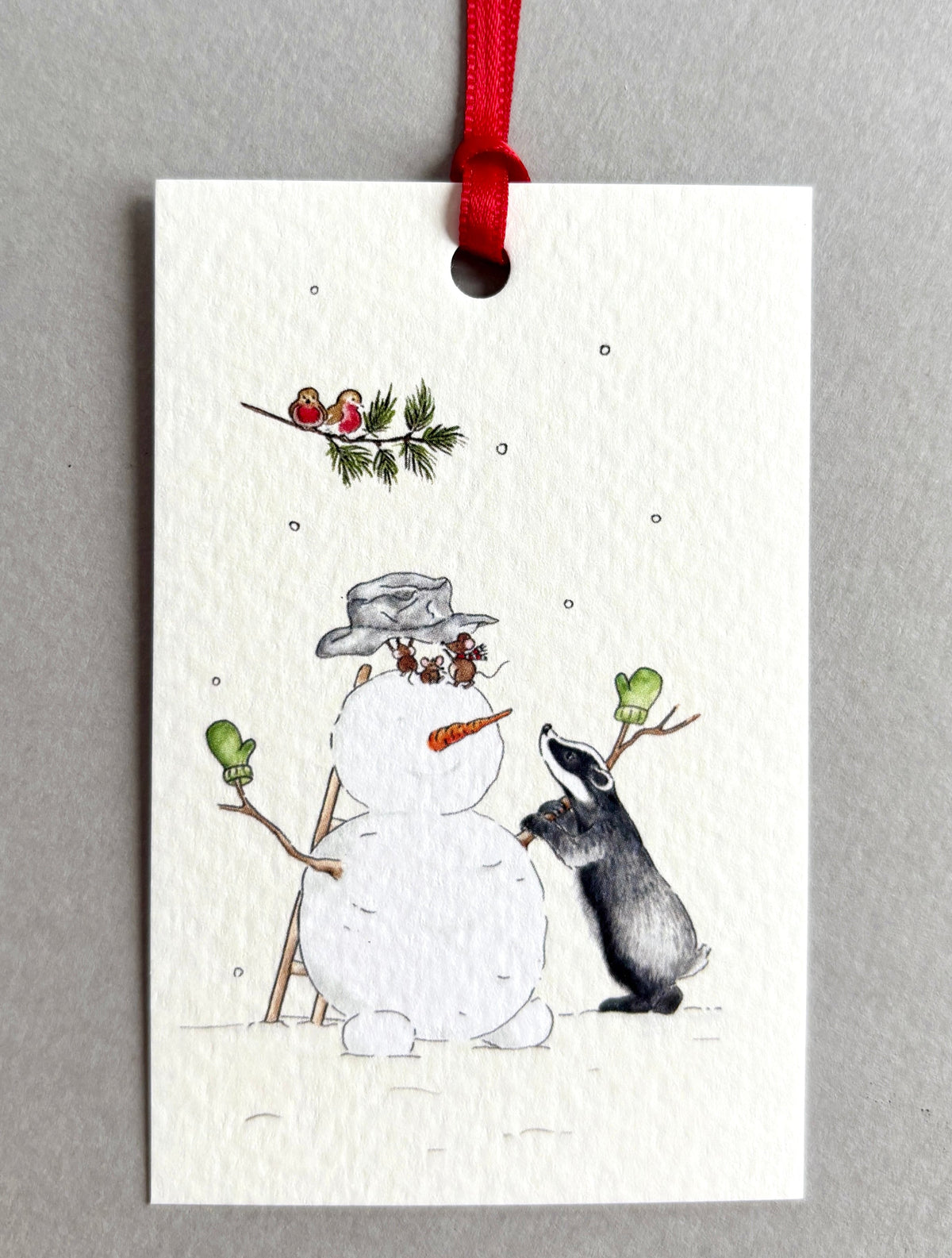 A rectangular Christmas gift tag with a red ribbon, featuring a watercolour illustration of a snowman and a badger under a fir tree branch with two robins. Coordinate with Snowman Wrapping Paper  