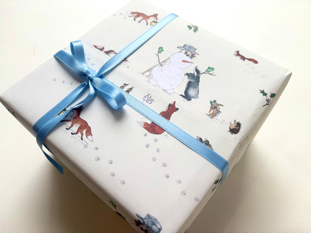 A gift box wrapped in cream wrapping paper featuring a whimsical woodland animal and snowman print, tied with a light blue satin ribbon and bow, on a light cream background.