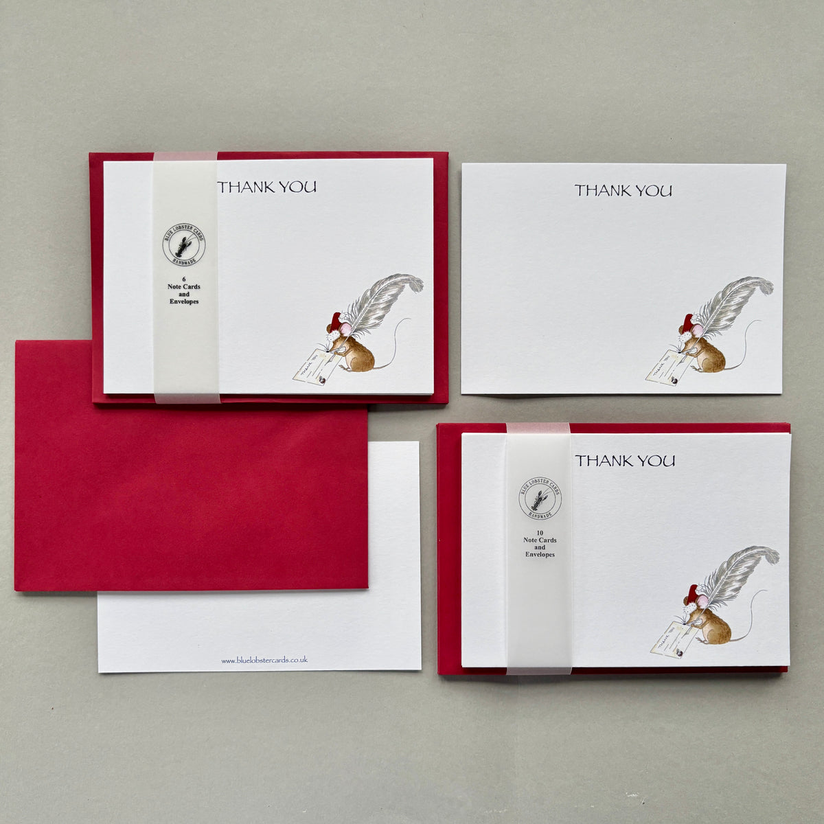 Sets of 6 and 10 thank-you postcards and matching envelopes arranged on a light gray surface. The postcards are white with translucent Blue Lobster Cards belly band, and a small illustration of a mouse holding a feather quill. Image shows single card. reverse with website details in very small print at the bottom of the card and Christmas red envelope