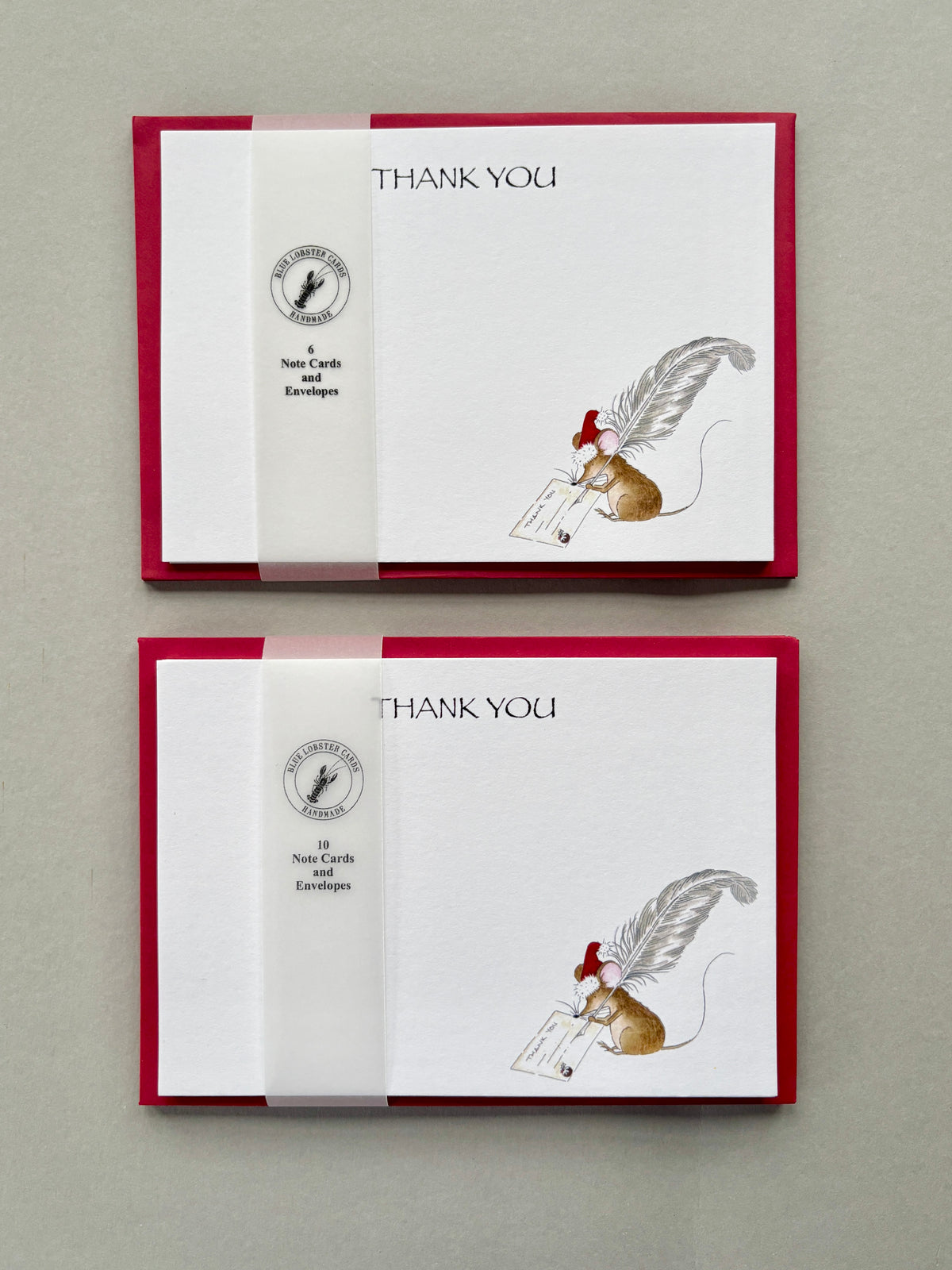 Two stacked Christmas thank-you card packs featuring a soft white matt textured card with Thank You text and an illustration of a mouse writing with a feather quill. Packs of 6 and 10 postcards wrapped in translucent Blue Lobster Cards belly band on a neutral gray background.