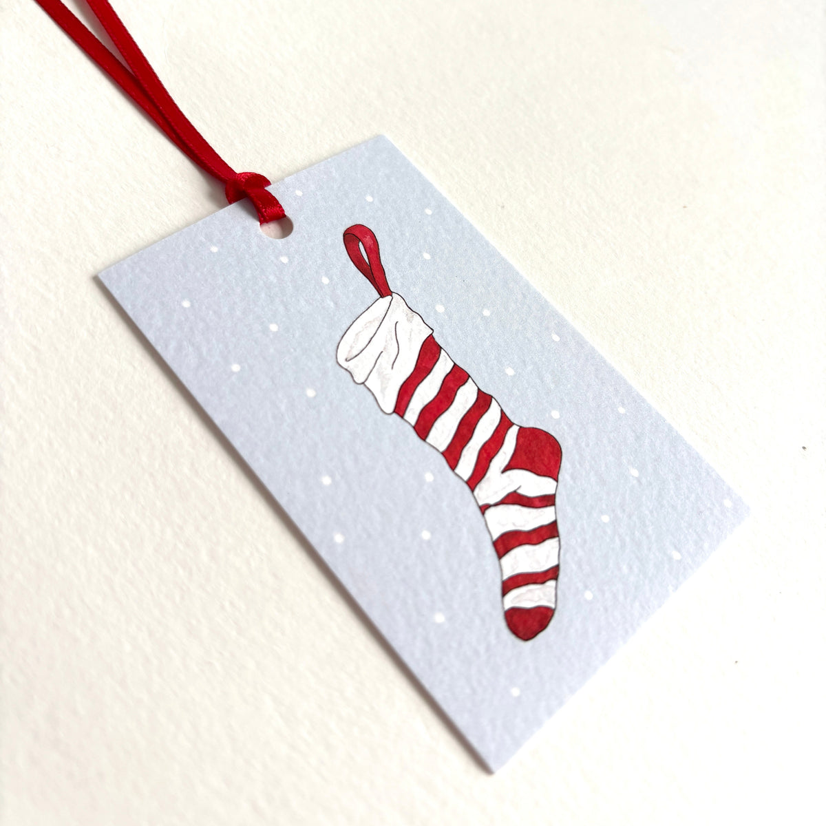 A rectangular light blue gift tag dotted with snow, with a watercolour illustration of a red-and-white striped Christmas stocking. Hand finished with a red hanging ribbon. Printed in UK onto 300gsm softly textured matt cardstock.