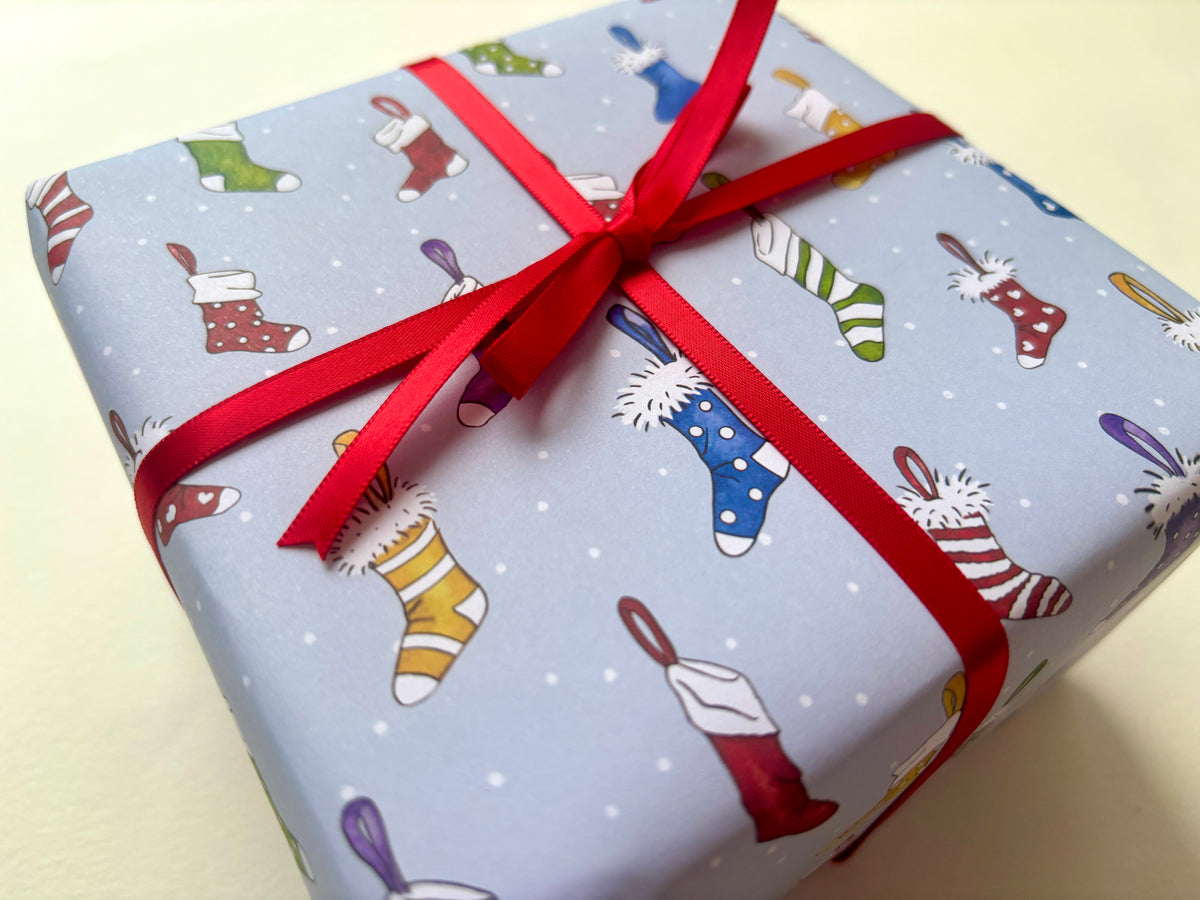 A sample gift box wrapped in light blue Christmas-themed paper with a pattern of colorful stockings and white snow dots, tied with a red ribbon bow.