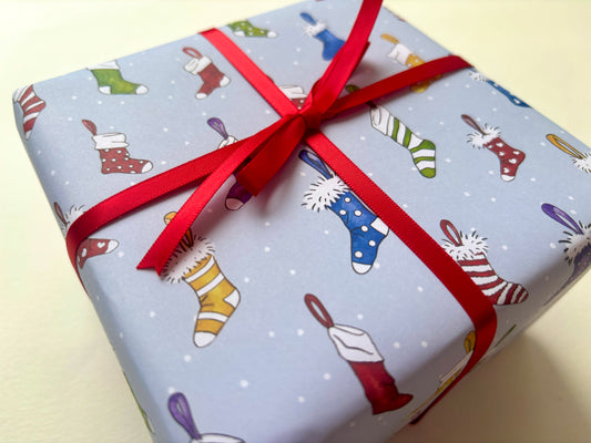 A sample gift box wrapped in light blue Christmas-themed paper with a pattern of colorful stockings and white snow dots, tied with a red ribbon bow.