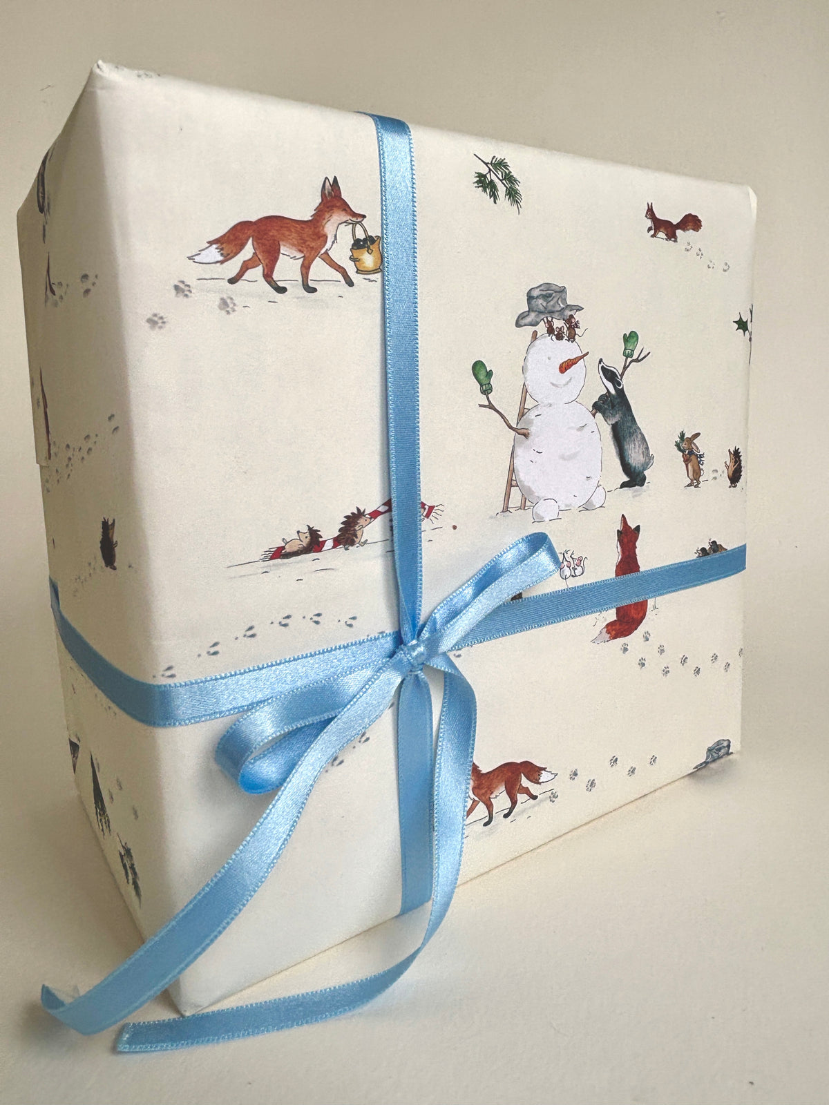 A gift box wrapped in Snowman Christmas wrapping paper featuring a winter woodland animals print of foxes, a snowman, and a dog, tied with a light blue satin ribbon against a cream background. 