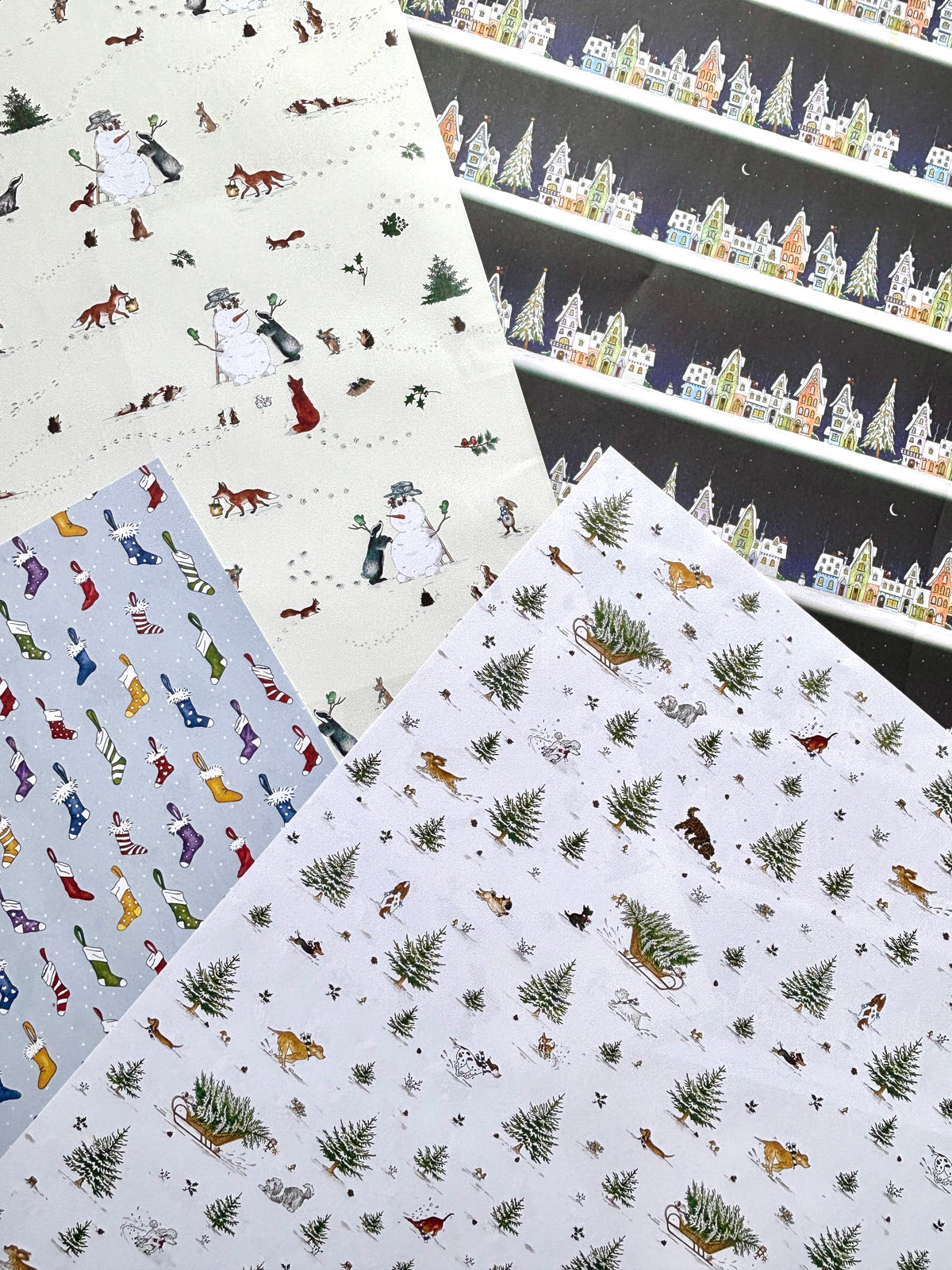 Four original watercolour designs of Christmas-themed wrapping paper. The designs include a snowy village on Christmas Eve with Father Christmas, snowmen with animals on a cream background, colorful, Christmas stockings on a light blue background with snow dots, lots of dogs playing in the snow amongst Christmas trees and sleighs on a white background.