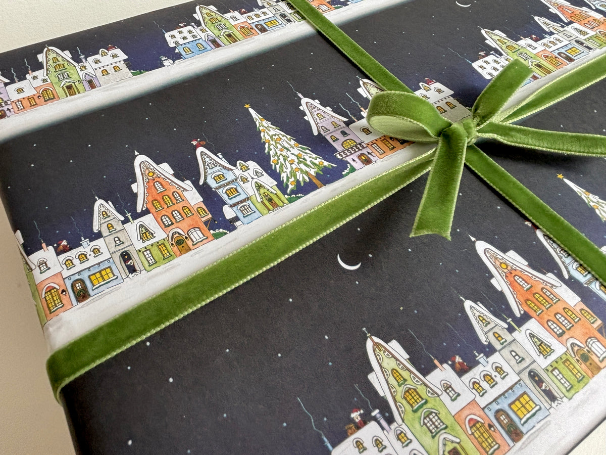 A close-up of Christmas wrapping paper with a snowy night sky, watercolour illustrated colourful houses and trees, finished with a sample green velvet ribbon tied in a bow.