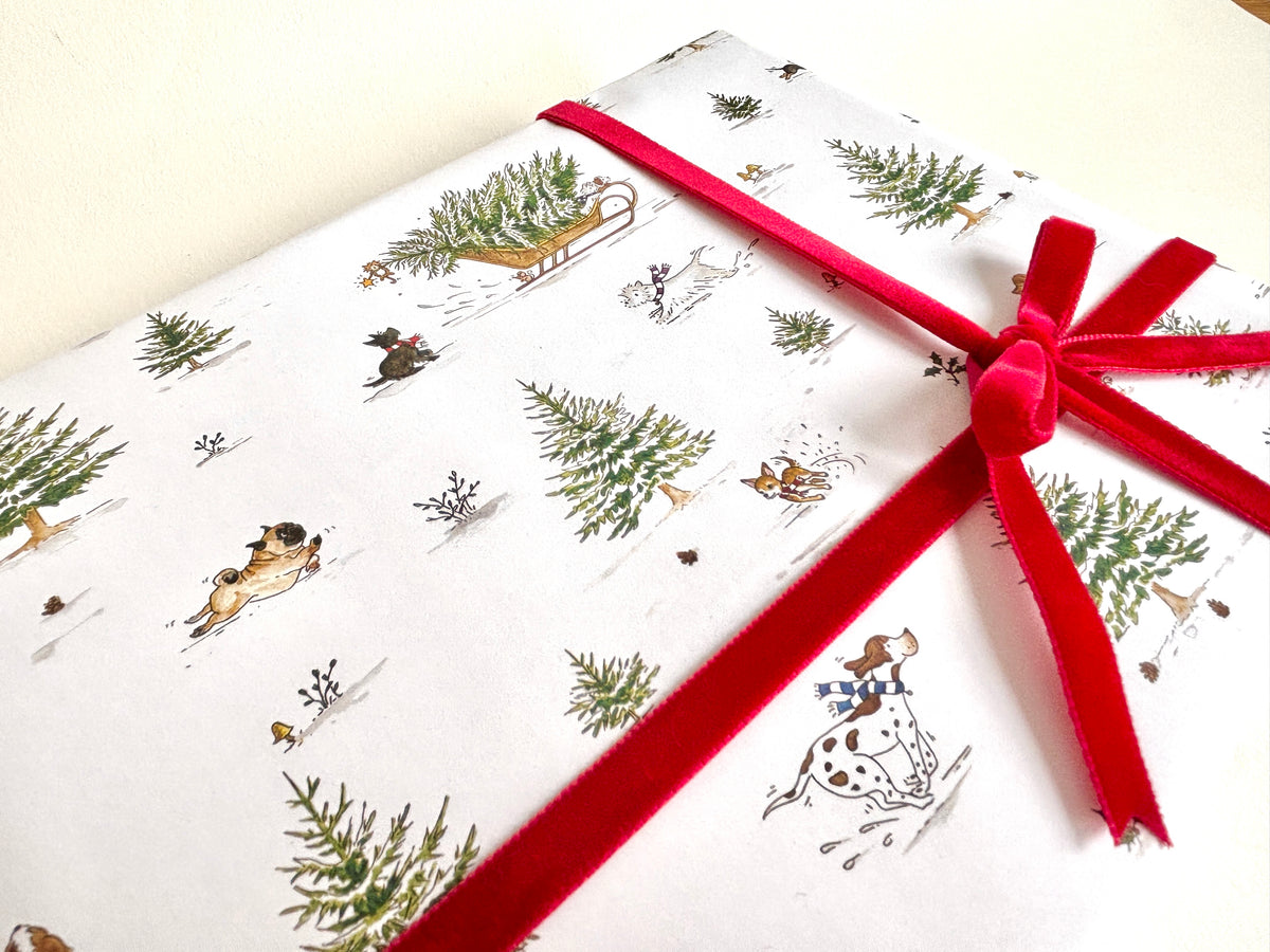 A close-up of Snow Dogs Christmas wrapping paper featuring a vintage-style winter scene with lots of dog breeds playing amongst Christmas trees. Here we see a pointer, pug, chihuahua, Scottish terrier etc. Gift box is tied with a bright red velvet ribbon - for display only - ribbon not available from Blue Lobster Cards. Matching gift tags available. 