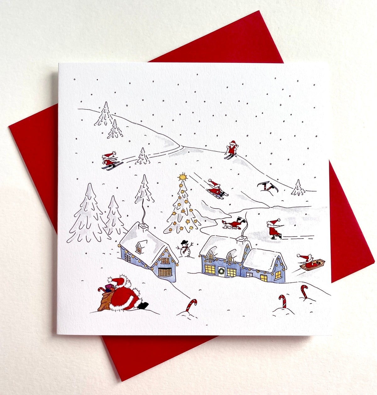 A square white greeting card showing a hand-drawn winter holiday snow scene, with Santa Claus, wtaching his helpers hae fun amidst snow-covered houses, trees, and sledders. Packs and single cards are supplied with red envelopes.