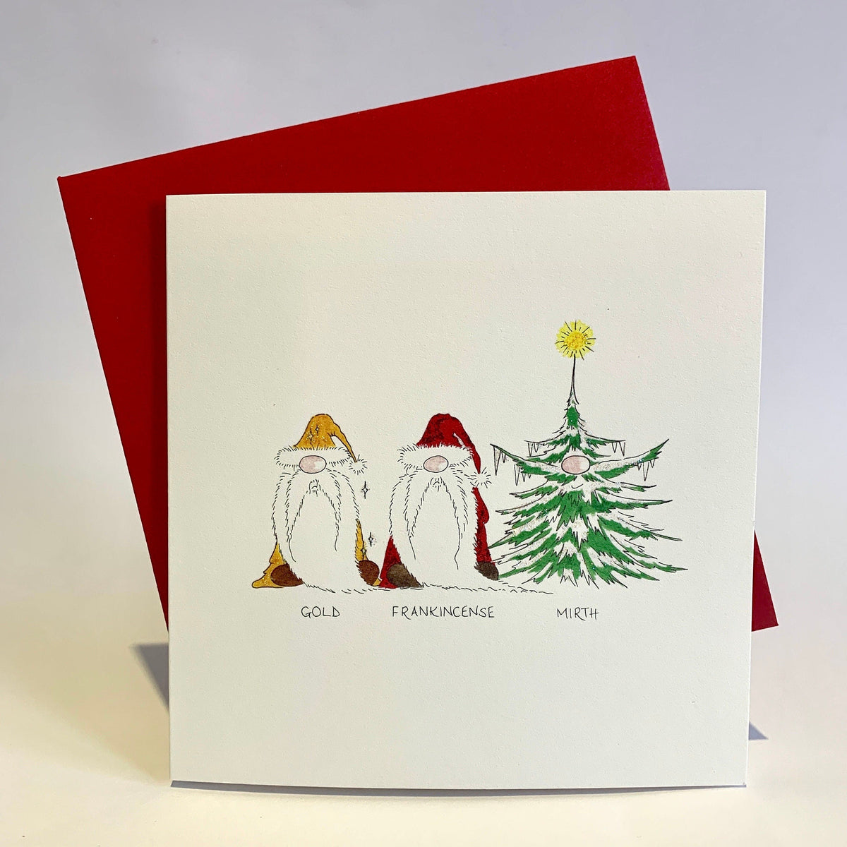 Funny Christmas card with a watercolour design featuring three gonks representing the three wise men, with 'Gold, Frankincense & Mirth' written on it. The card is held against a red envelope on a white background.