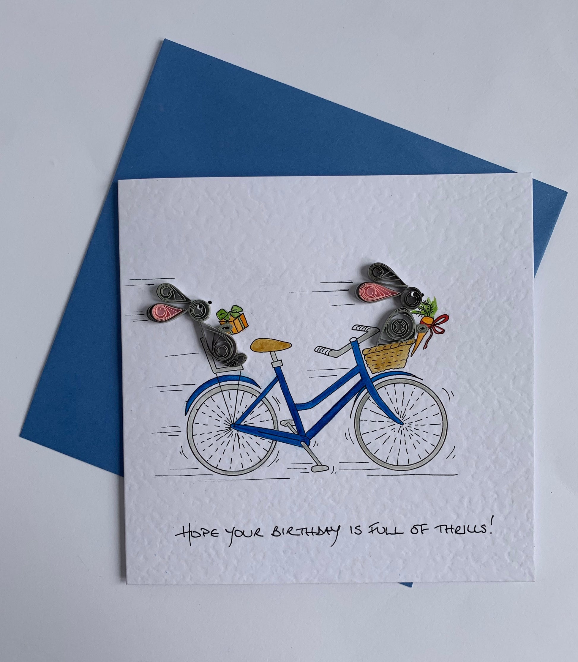 Hand painted bicycle bunny card shoing 3D quilled paper rabbits with gifts displayed on hammered card with blue envelope