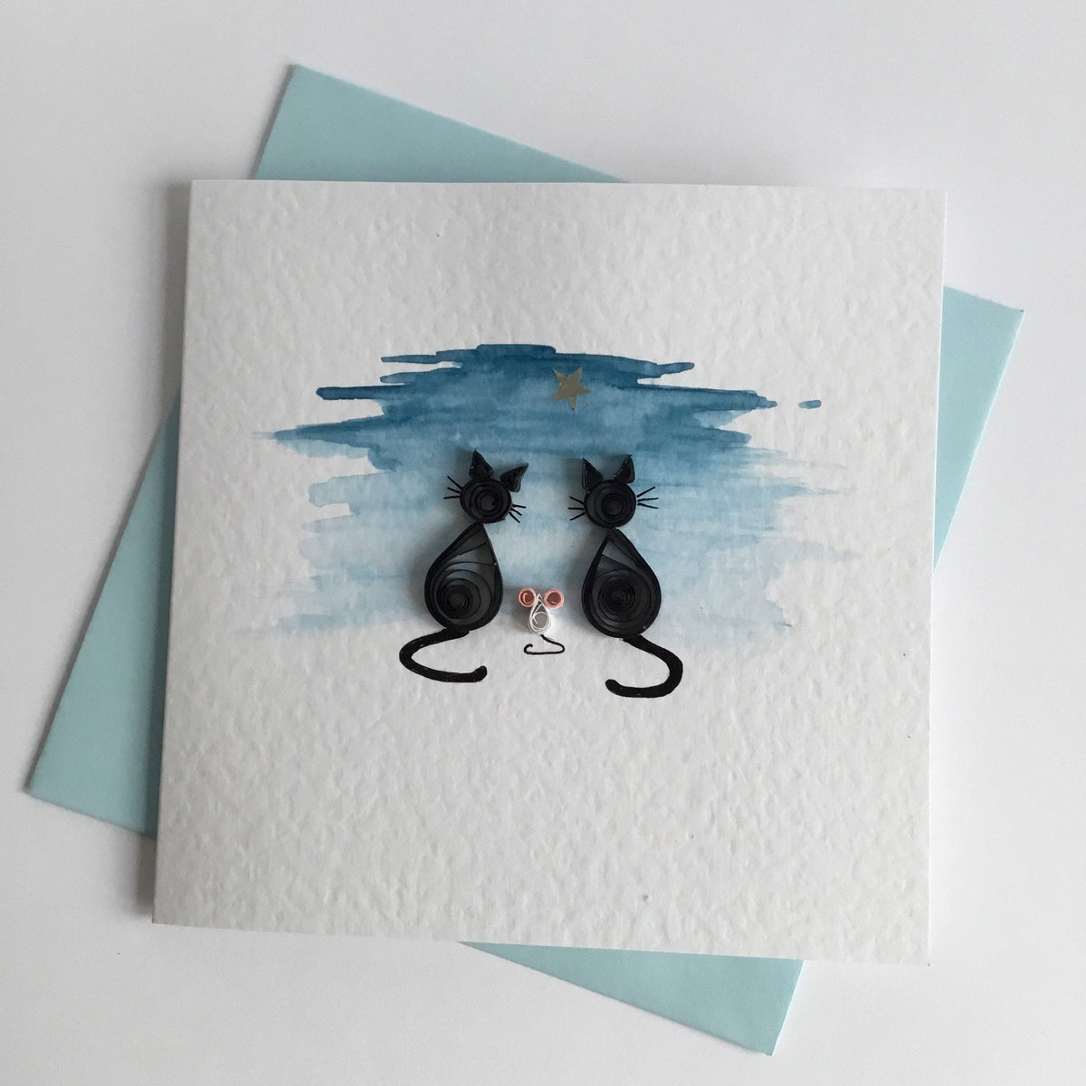 Handmade card with a watercolour illustration of two 3D quilled black cats and a white mouse looking out at  a silver star, set against a hand painted dusk blue sky. The card has a blank interior for a personal message and comes with a blue envelope.