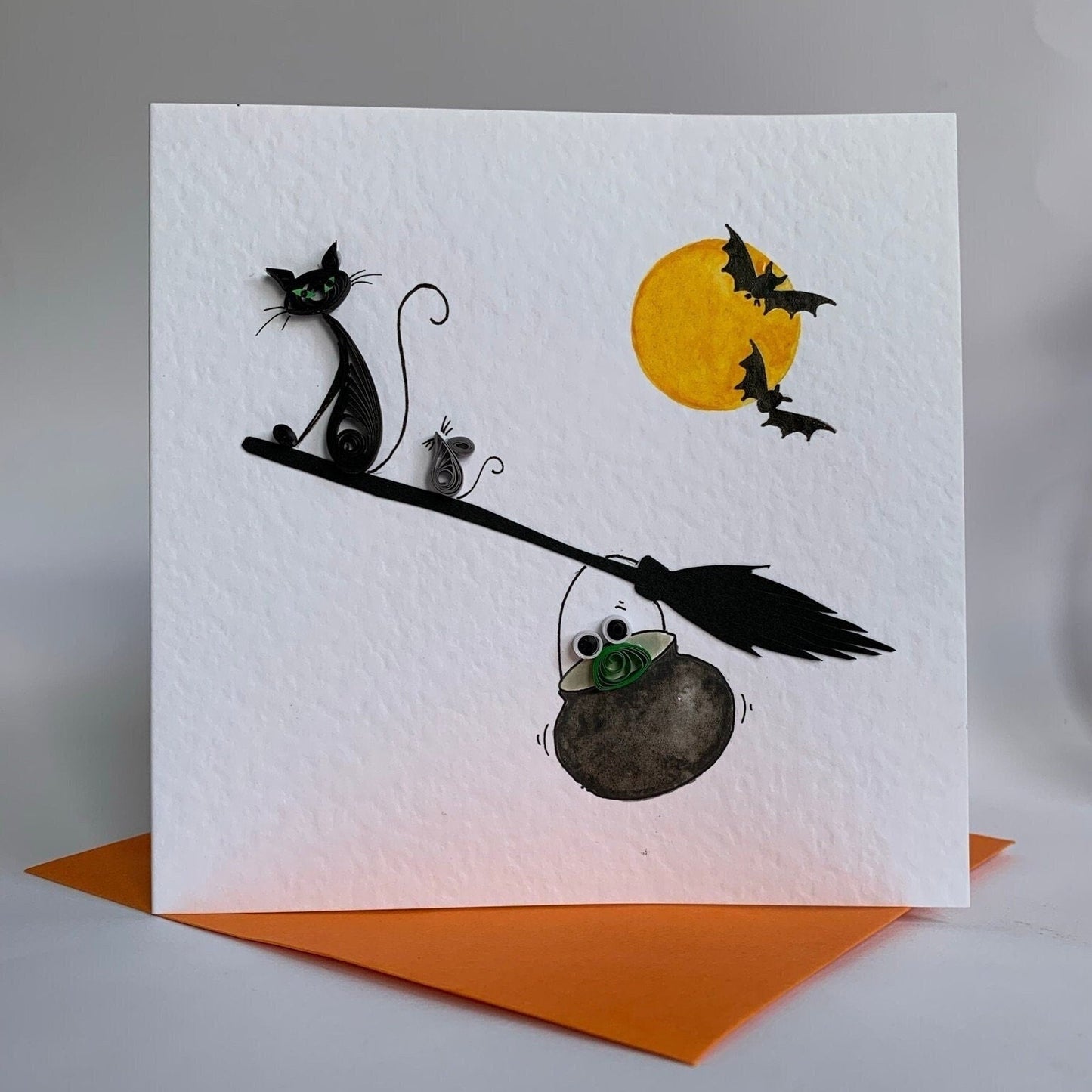 Handmade Halloween greeting card with quilled witch's black cat, mouse, and frog on a broomstick, with hand-painted orange moon and bat silhouettes. The card is displayed with an orange envelope.