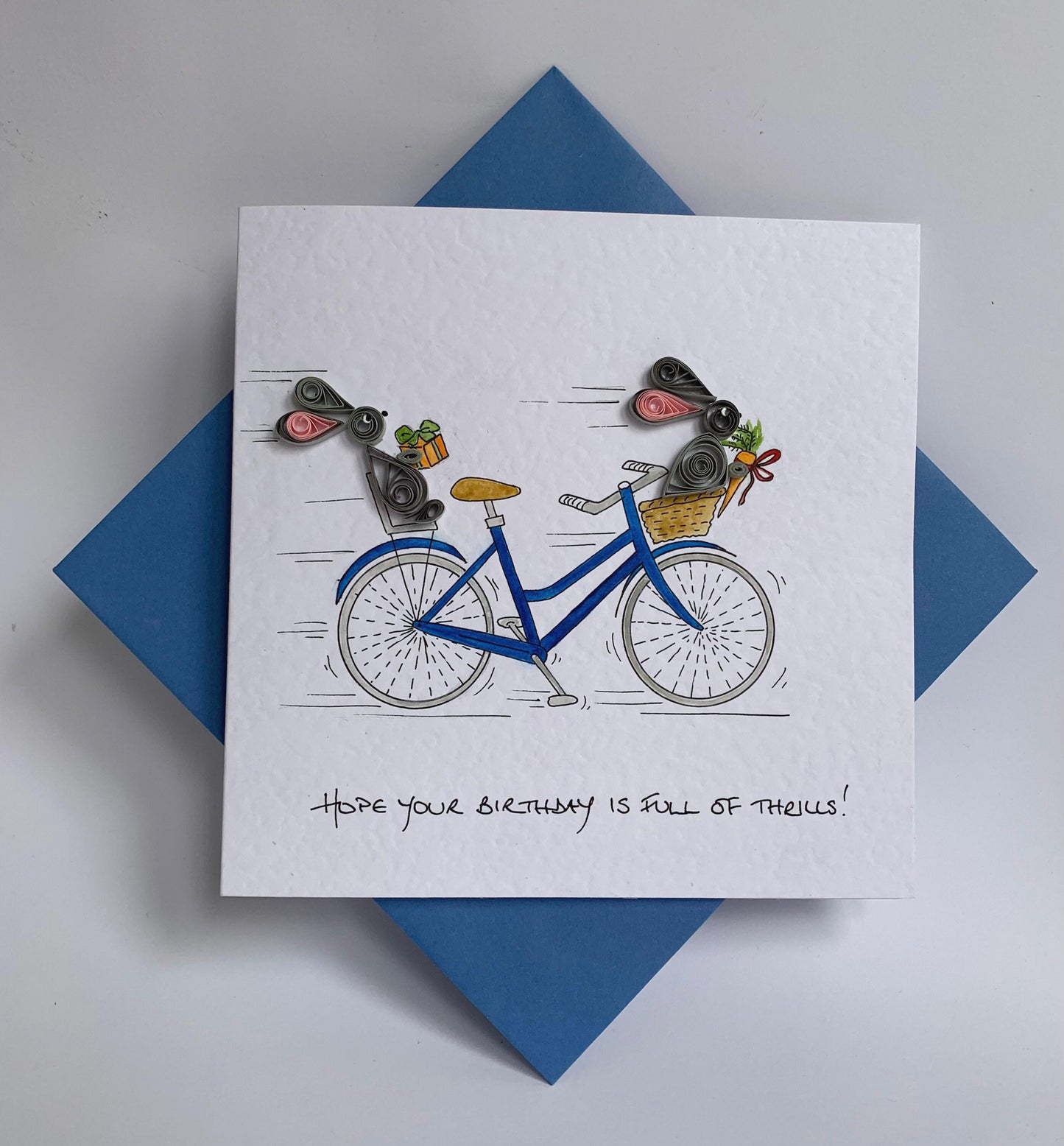 Handmade quilled birthday card featuring two quilled 3D paper rabbits  racing on a hand painted blue bicycle. shows matching blue envelope