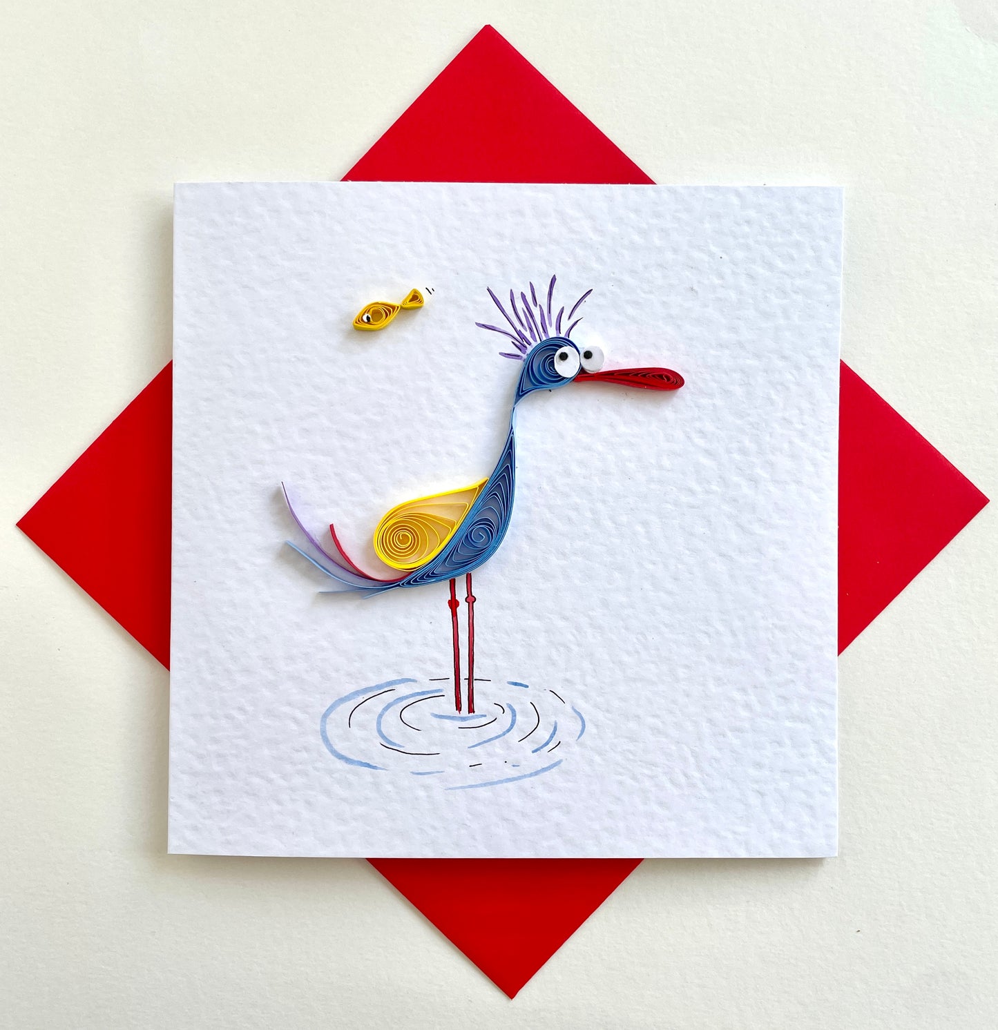 Quilled greetings card of a bird with a purple tuft on its head and a red beak, standing on one leg, with a fish jumping above it. The card is blank inside and includes a matching red envelope.