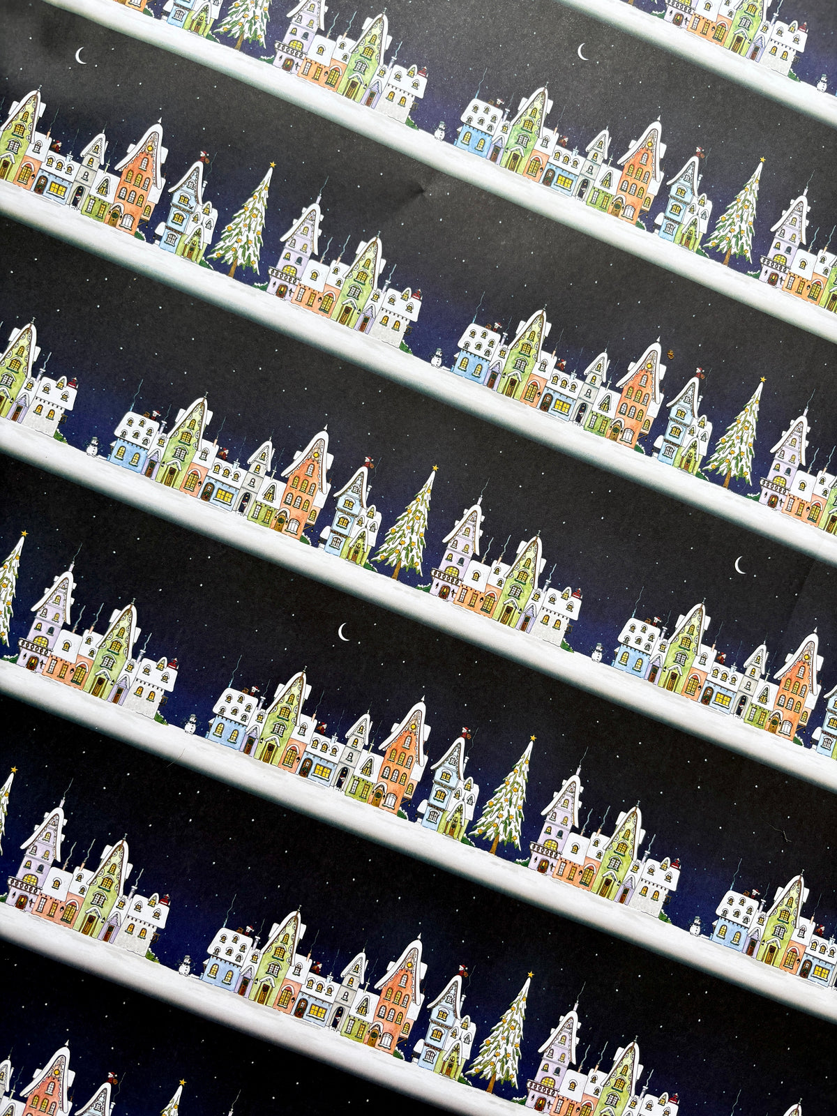 A close-up of Snowy Christmas Eve wrapping paper showing watercolour illustration with repeating pattern of a winter village scene with Father Christmas making his way along the rooftops of colourful houses, trees with snow, stars, and crescent moons on a dark navy background.