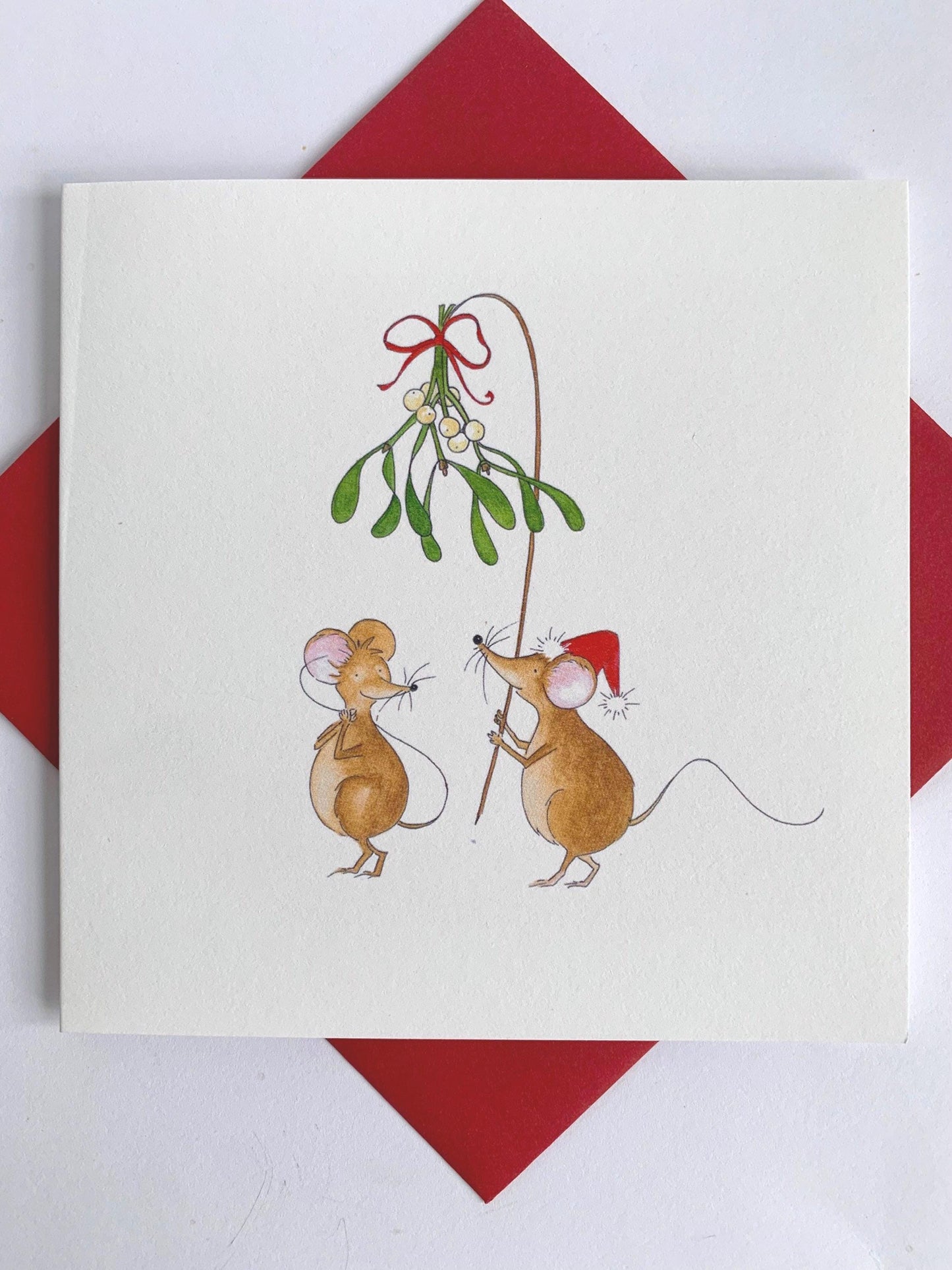 Square charity Christmas card set against coordinating red envelope shows two cute mice, one with a Christmas hat and holding aloft a branch of mistloe tied with red ribbon, hoping for a Christmas kiss. Designed and printed in the UK from original watercolour artwork