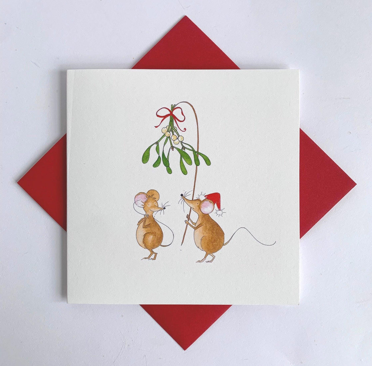 Charming watercolour illustration of a mouse with mistleote tied with festive ribbon hld above another mouse, hoping for a Christmas kiss. Set against a red envelope