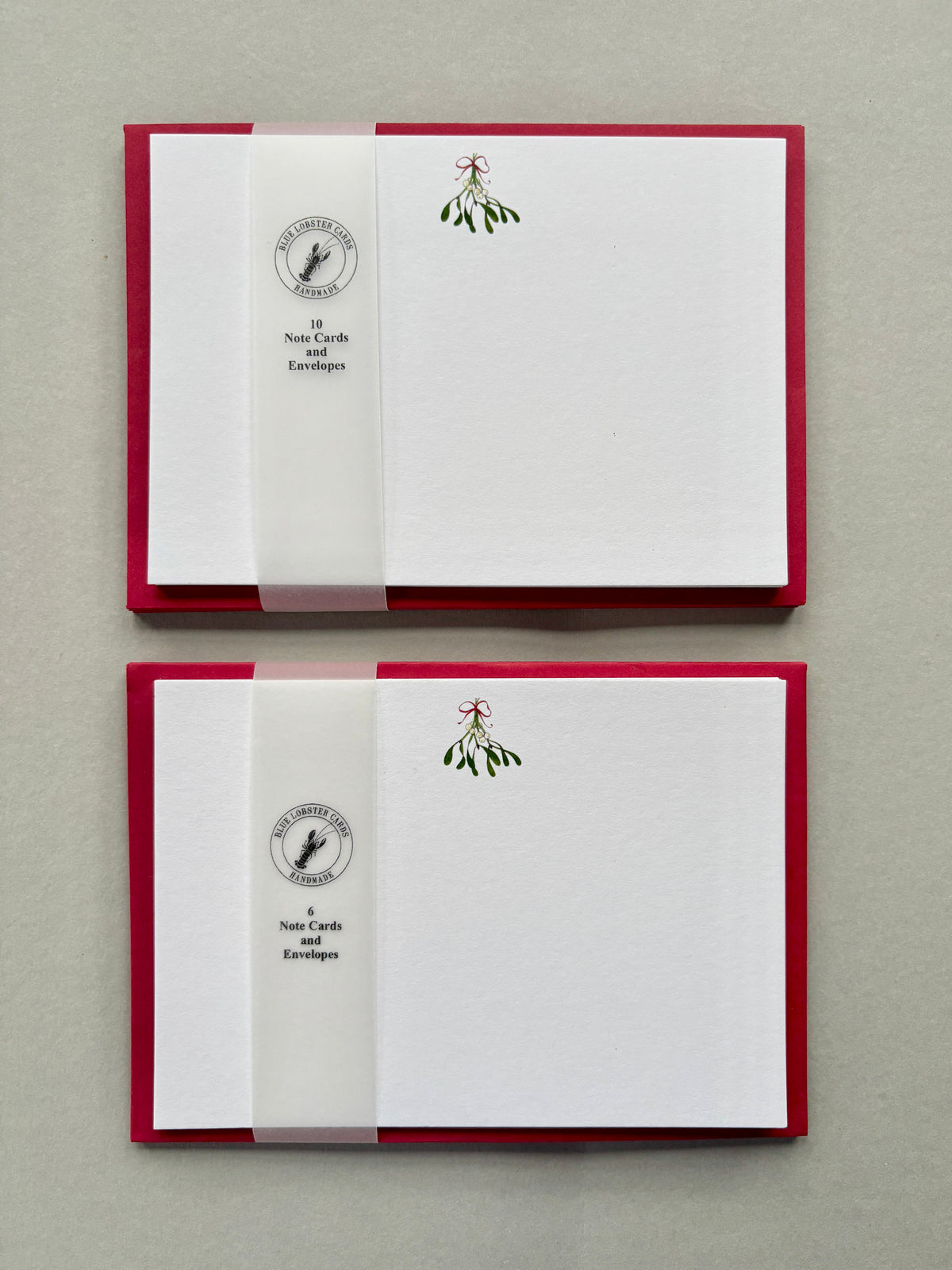 Two stationery sets of Christmas notecards with red envelopes featuring a mistletoe illustration, set against a neutral gray background. packaged in recyclable transulcent belly band
