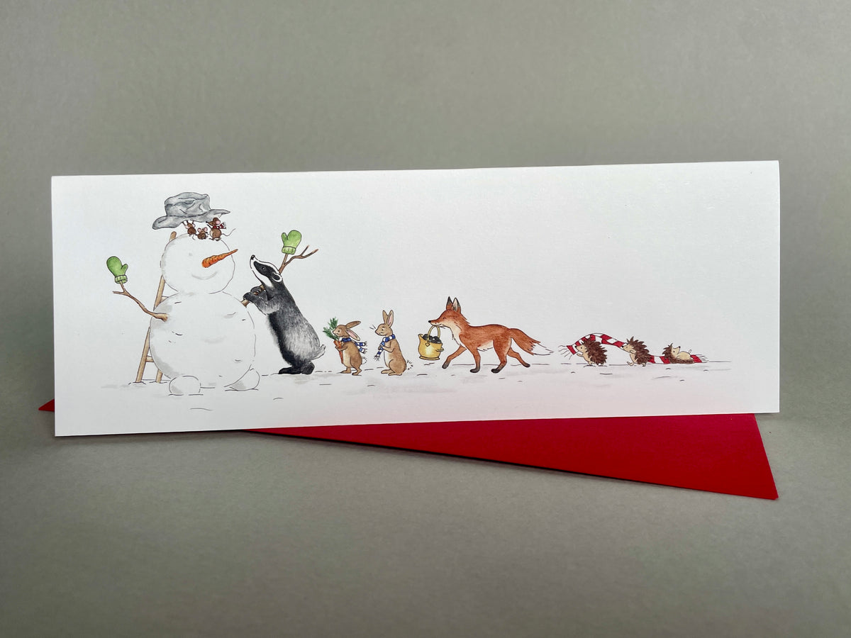 Full woodland animals snowman scene on an upright Christmas card - long slim shape with a red envelope on a grey surface