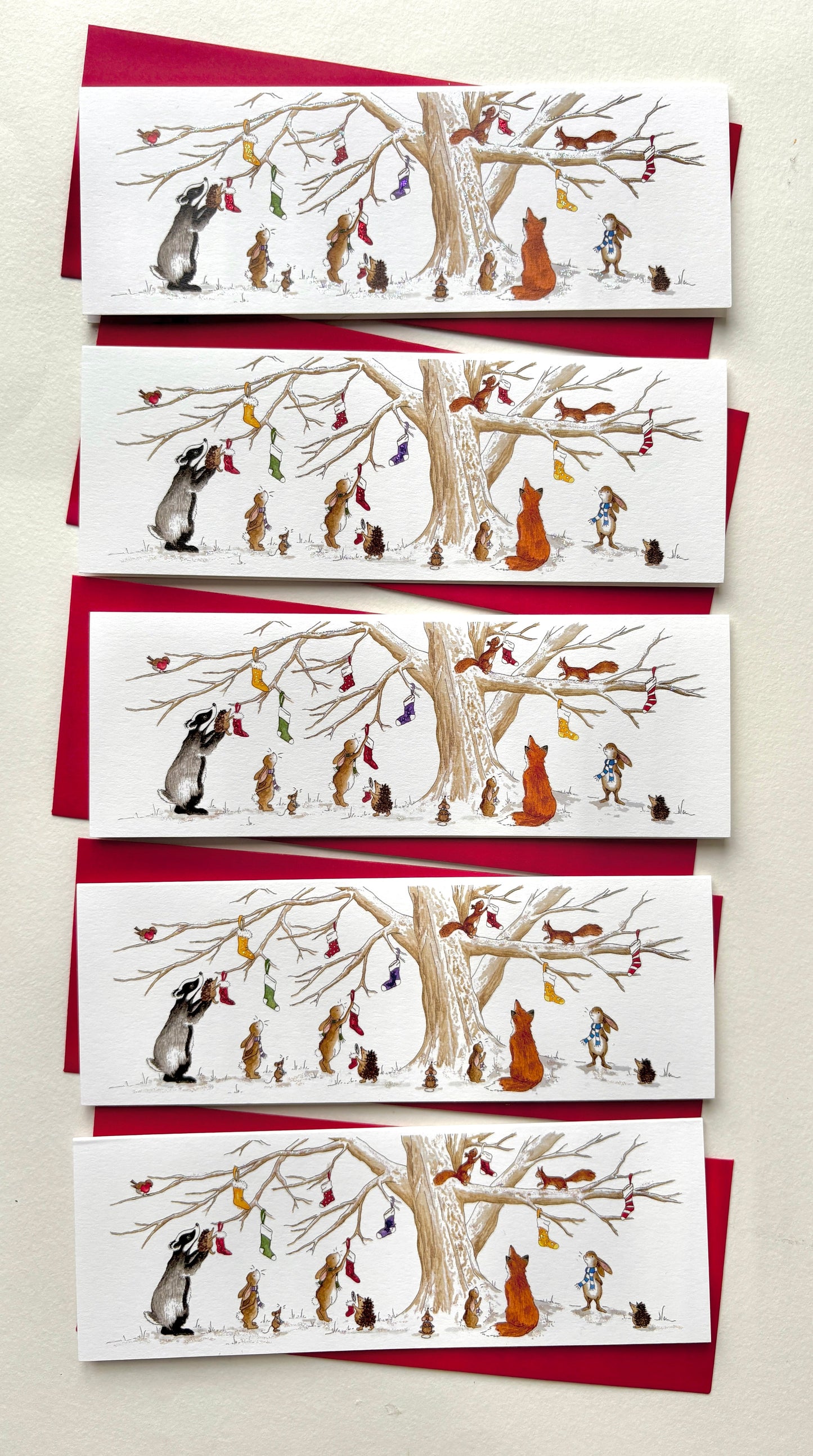 PAck of 5 cards - same design - featuring a badger, rabbits / bunnies, fox, squirrels and hedgehogs with colourful scarves hanging stockings on a snowy winter tree. Glitter version is show - non-glitter available.