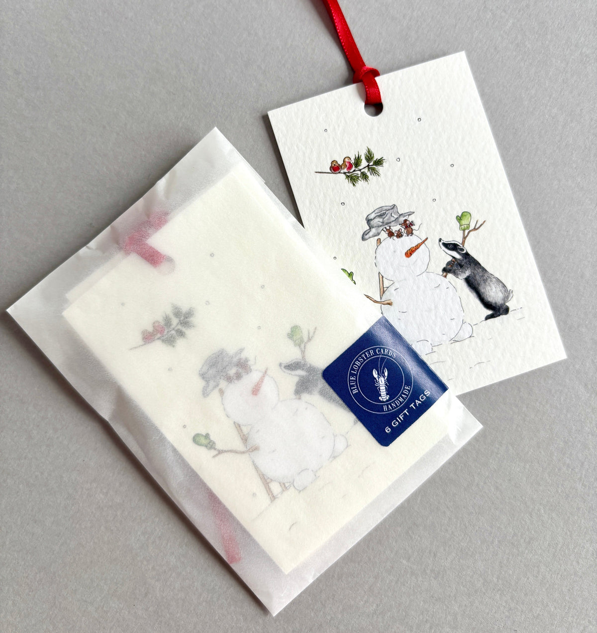 Pack of 6 Christmas gift tags with watercolour illustration of a snowman, a rabbit, and robins perched on a fir branch. Printed in UK in fine quality textured cardstock and hand finished with red satin ribbon. Tags are presented in a recyclable glassine bag and Blue Lobster Cards branding . 
