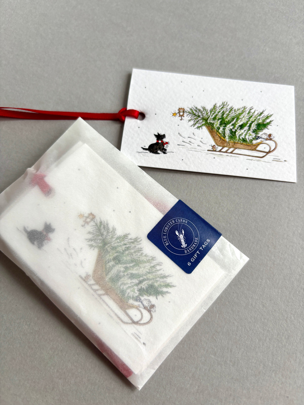 Packaging for a set of 6 Christmas gift tags with an original watercolour illustration of a sleigh carrying a Christmas tree, with a Scottie dog looking up at a cat dangling from a branch. 6 tags, hand finished with red ribbon are packaged in recyclable glassine sleeve with Blue Lobster branding. 