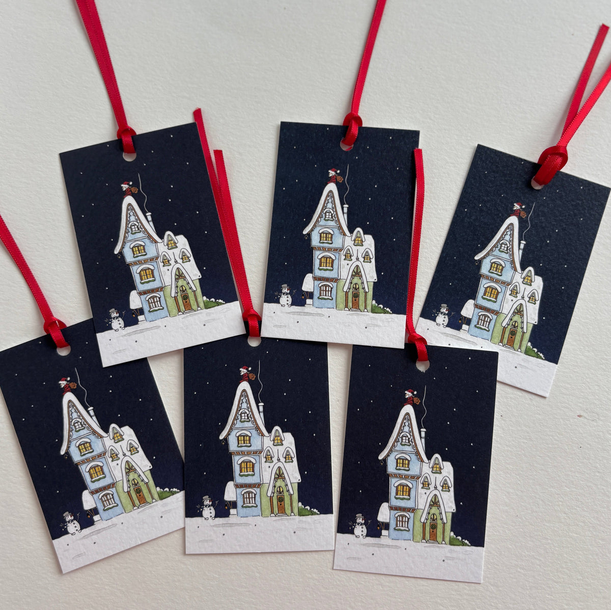 Six Christmas gift tags with red hanging ribbons, featuring an illustrated gingerbread house with Father Christmas and snowman on a dark navy snowy night sky background.