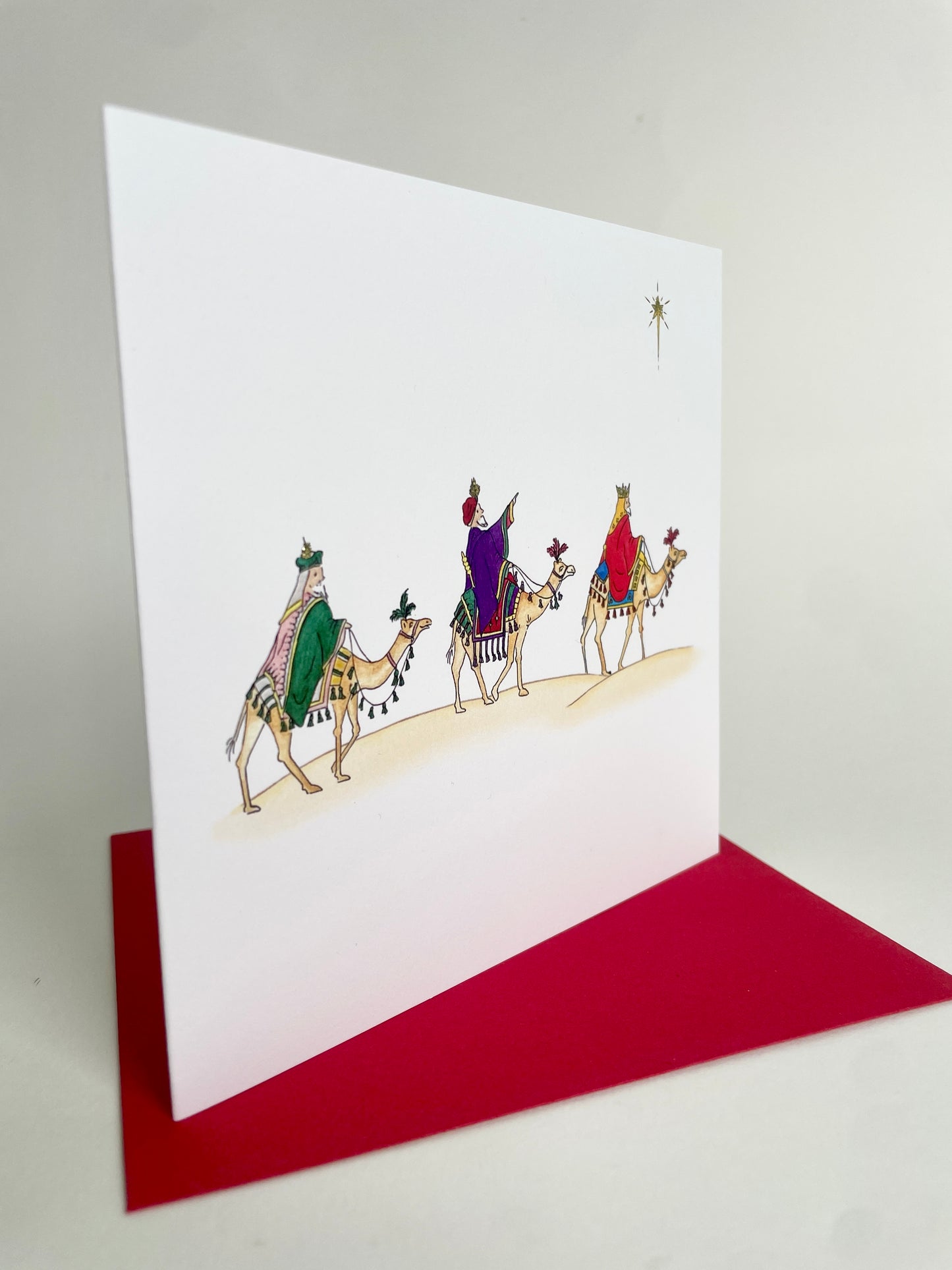Angled image of three kings Christmas card with watercolour illustration of three wise men on camels, featuring a red envelope.