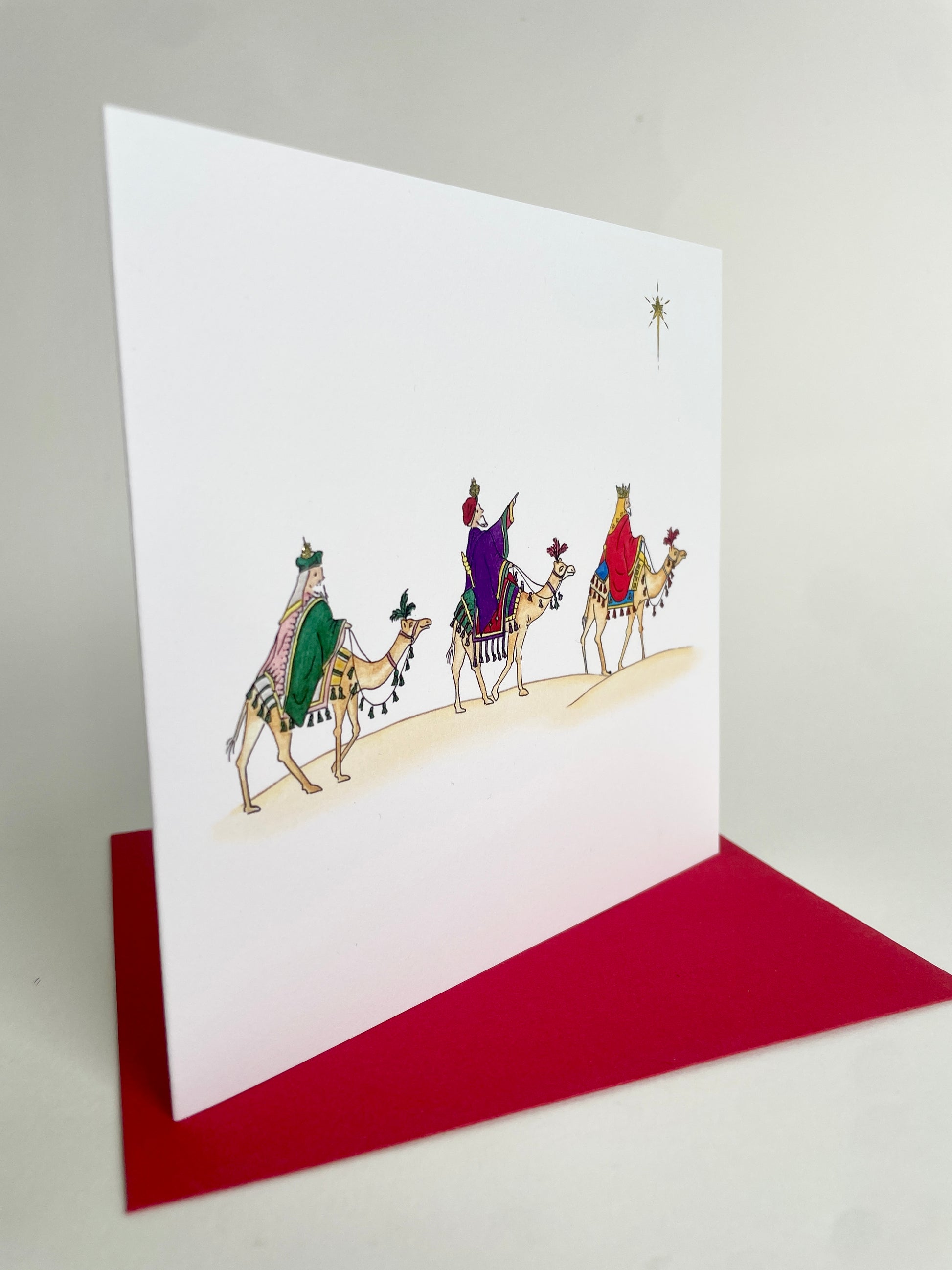 Angled image of three kings Christmas card with watercolour illustration of three wise men on camels, featuring a red envelope.