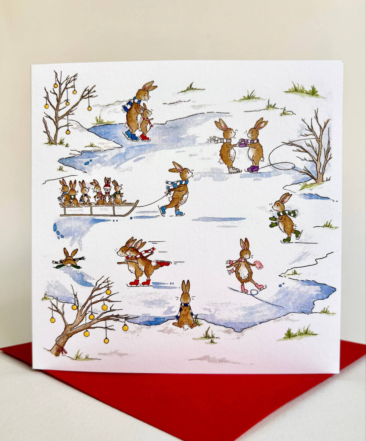 Charming winter scene of a frozen lake crowded with lively rabbits, skating and sledging. Design created by Blue Lobster Cards and printed in UK using fine qualtiy 300gsm FSC cardstock. Square card design rests on coordinating red envelope.