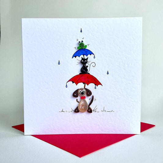 A hand painted and quilled greeting card with a  brown dog with a wagging tail, a black cat on top of a red umbrella, and a blue umbrella with a green frog.