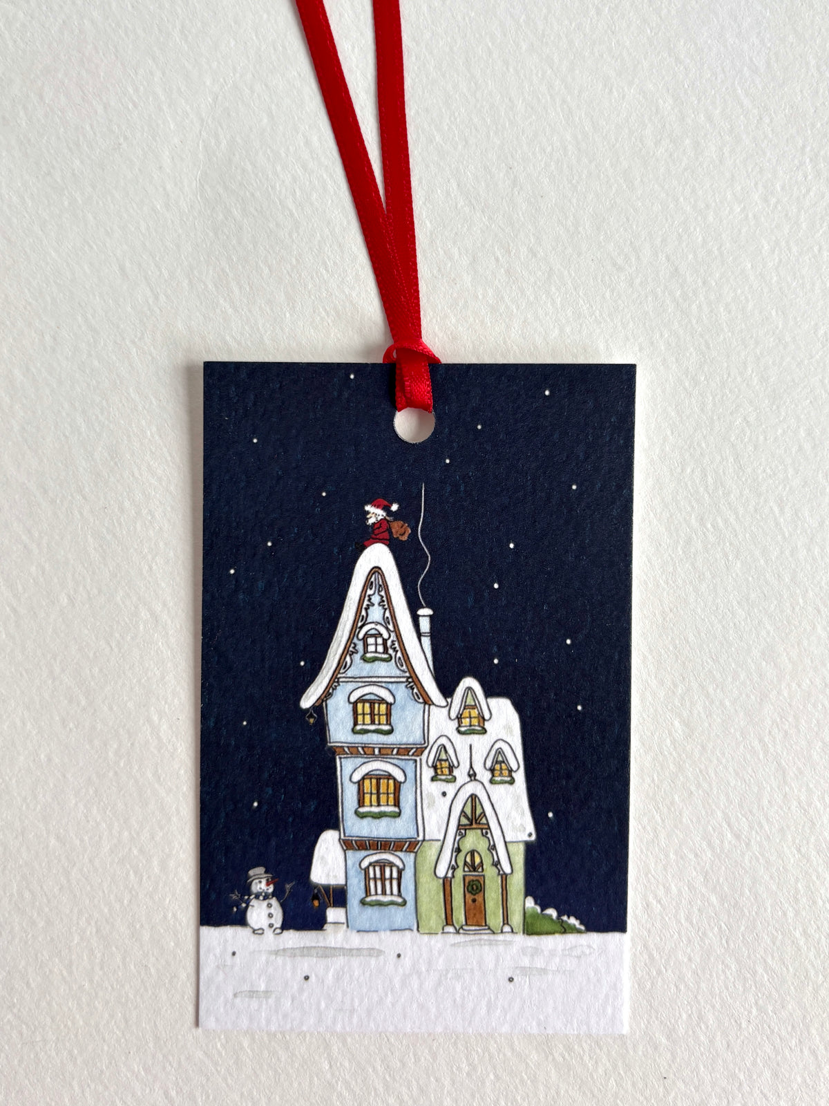 A rectangular Christmas ornament or gift tag with a red hanging ribbon, featuring a hand-drawn winter house scene under a dark navy night sky. The illustration shows snow-covered rooftops, glowing windows, a small snowman, and Father Christmas on top of the house.