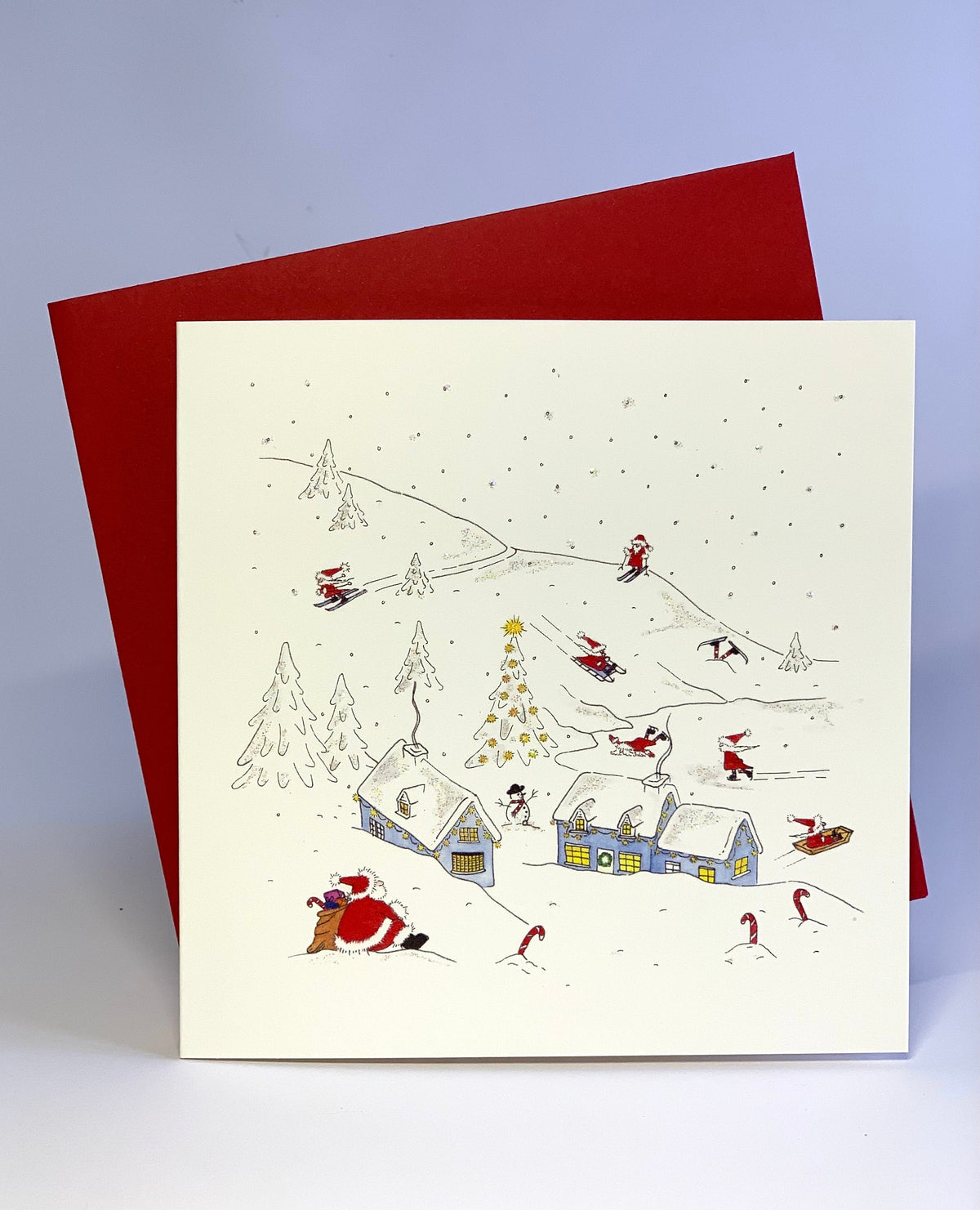 Christmas card showing Father Christmas looking at the Christmas elves skiing and sledging among snow-covered cottages and fir trees, with a red envelope behind