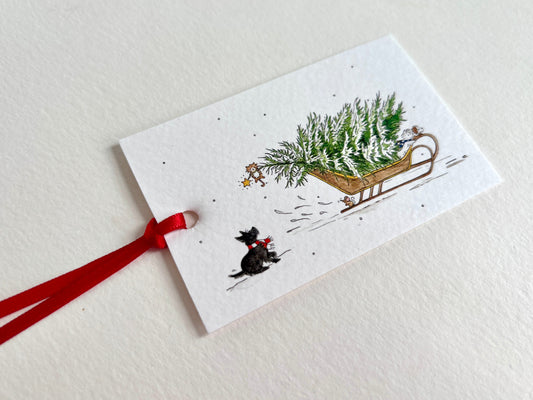 Rectangle gift tag hand finished with a red ribbon, featuring a whimsical illustration of a little black dog with striped scarf looking at a cat dangling from a snow covered Christmas tree in a sledge. 