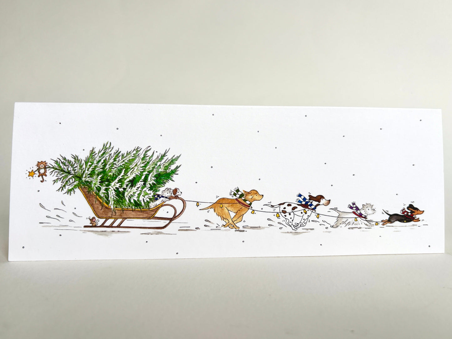Fun dog charity Christmas card featuring an original watercolour illustration of dogs pulling a Christmas tree through the snow on a sleigh