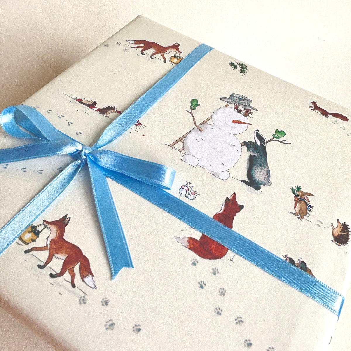 A gift box wrapped in cream-colored wrapping paper featuring a whimsical winter woodland print with a snowman, foxes, rabbits, and paw prints, tied with a light blue satin ribbon.