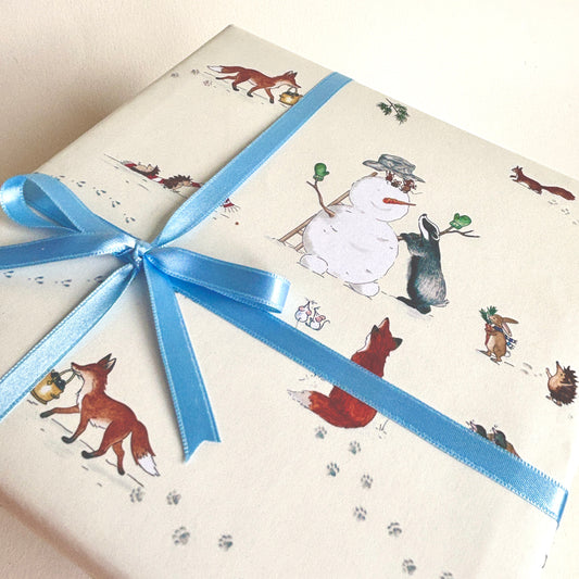 A gift box wrapped in cream-colored wrapping paper featuring a whimsical winter woodland print with a snowman, foxes, rabbits, and paw prints, tied with a light blue satin ribbon.