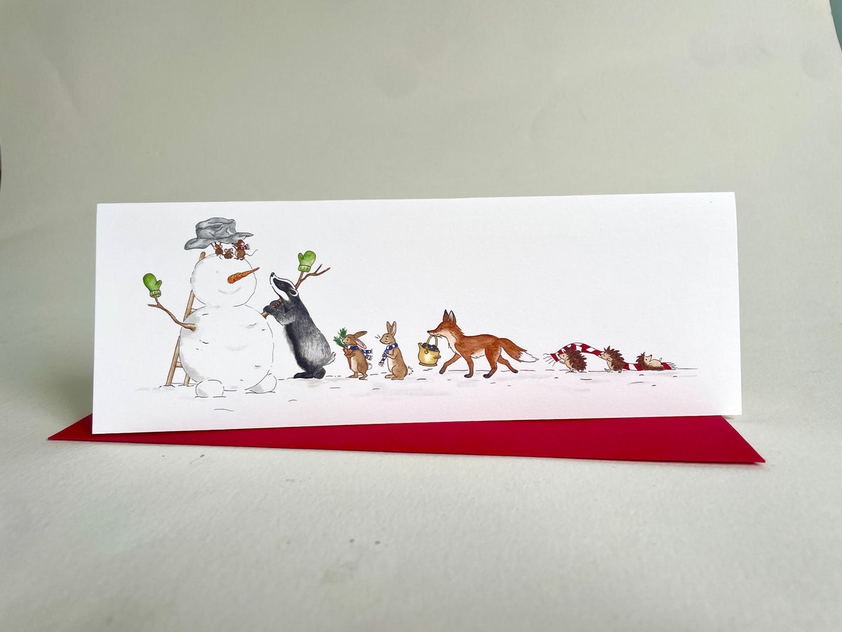 Woodland animals snowman Christmas card standing upright above a red envelope on a light background