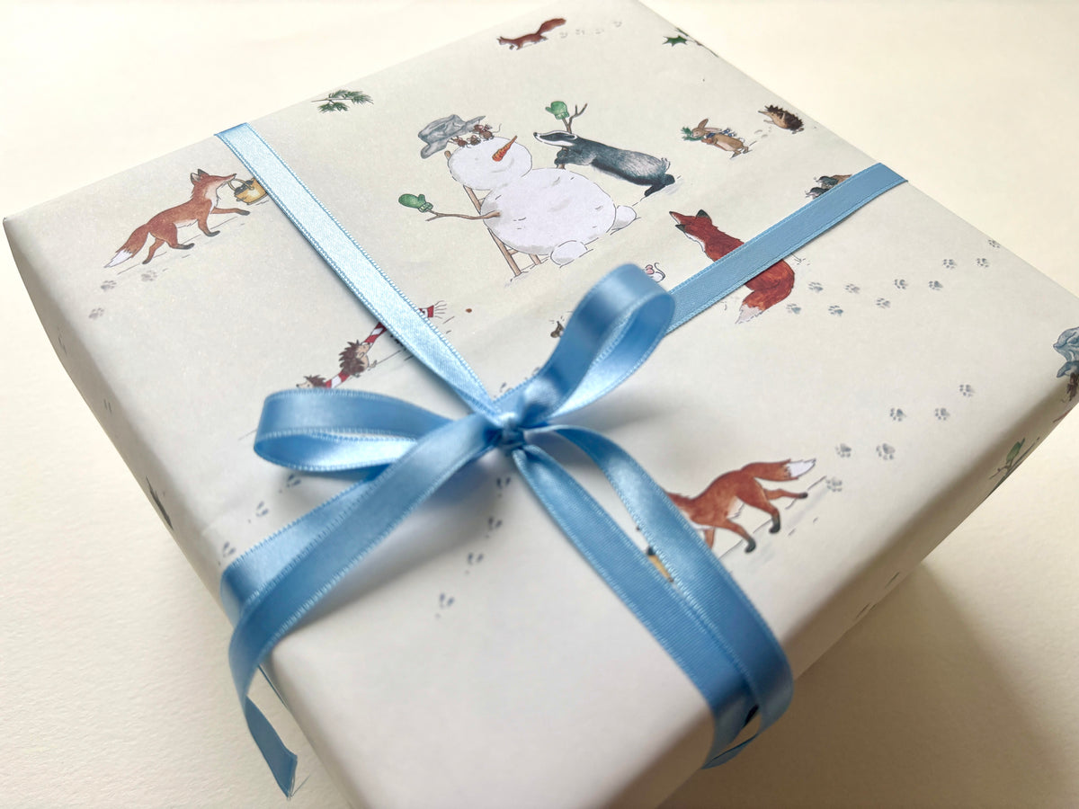 A gift box wrapped in attractive snowman wrap featuring British winter animals - foxes, badgers, hedgehogs, mice and bunnies / rabbits. Parcel is tied with a light blue satin ribbon in a bow (ribbon is not available to order and for decorative purposes only) 