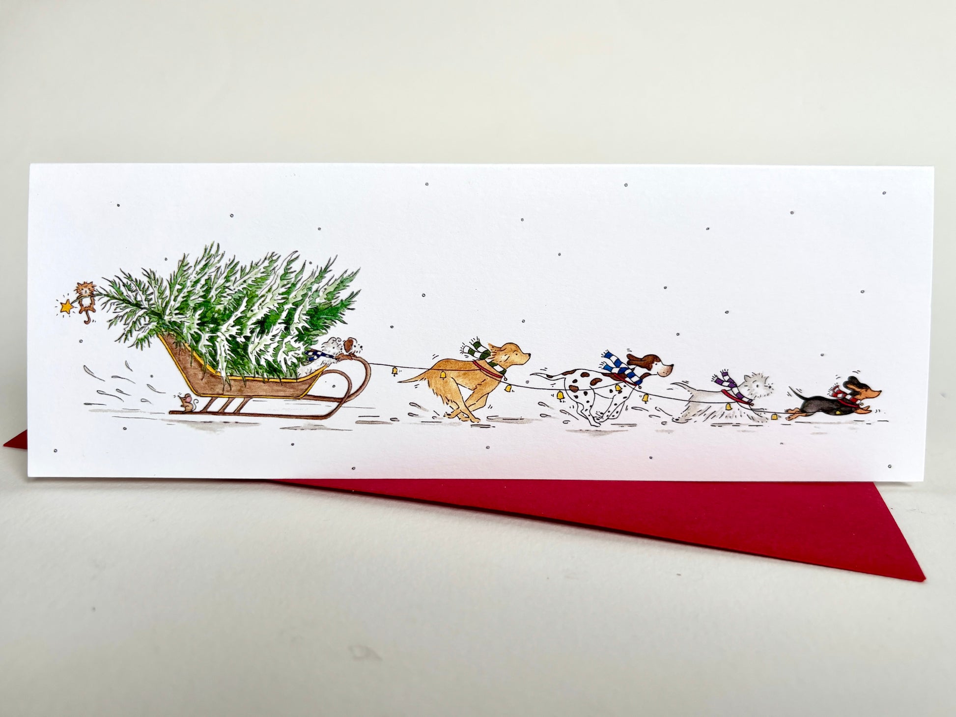Long slim charity Christmas card featuring four dog breeds - labrador retrieber, pointer, westie and dachshund with colourful striped scarves pulling a Christmas tree in falling snow.