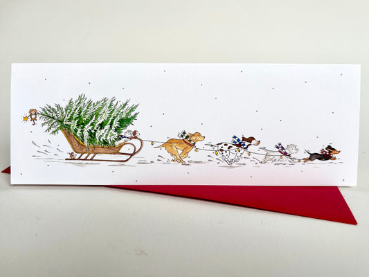 Long slim charity Christmas card featuring four dog breeds - labrador retrieber, pointer, westie and dachshund with colourful striped scarves pulling a Christmas tree in falling snow.
